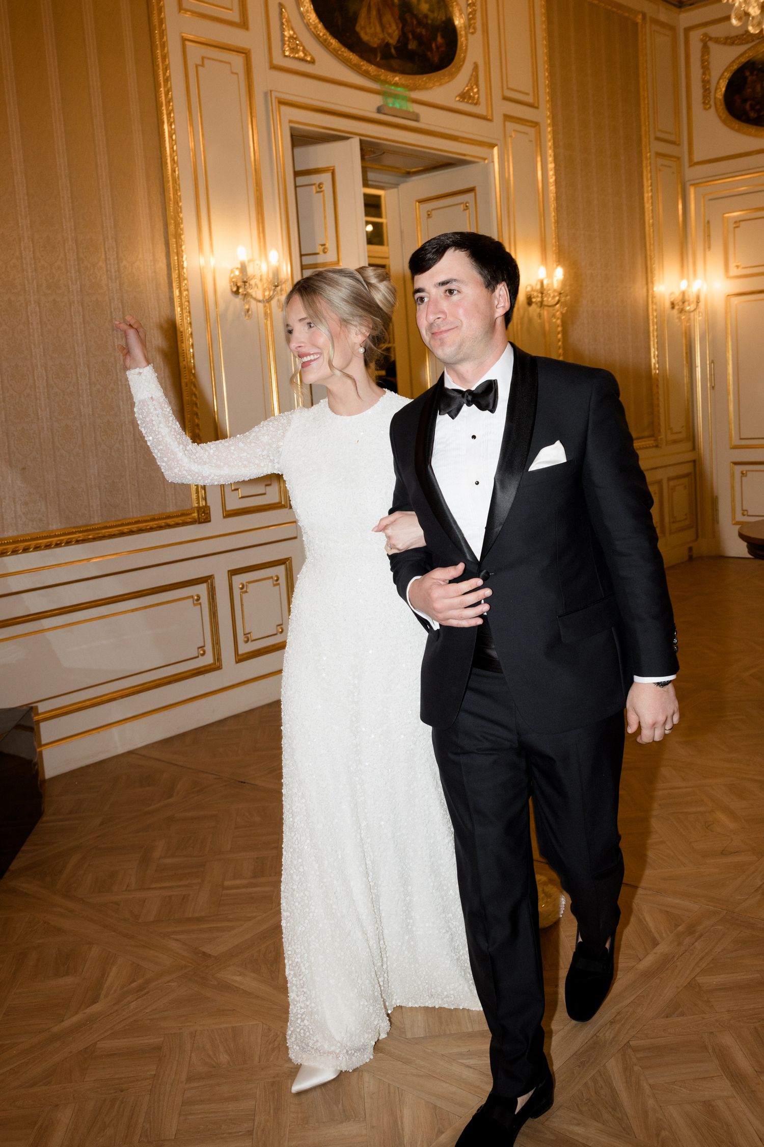 A couple in formal wedding attire walking arm in arm through an elegant ballroom with ornate gold walls and parquet flooring.