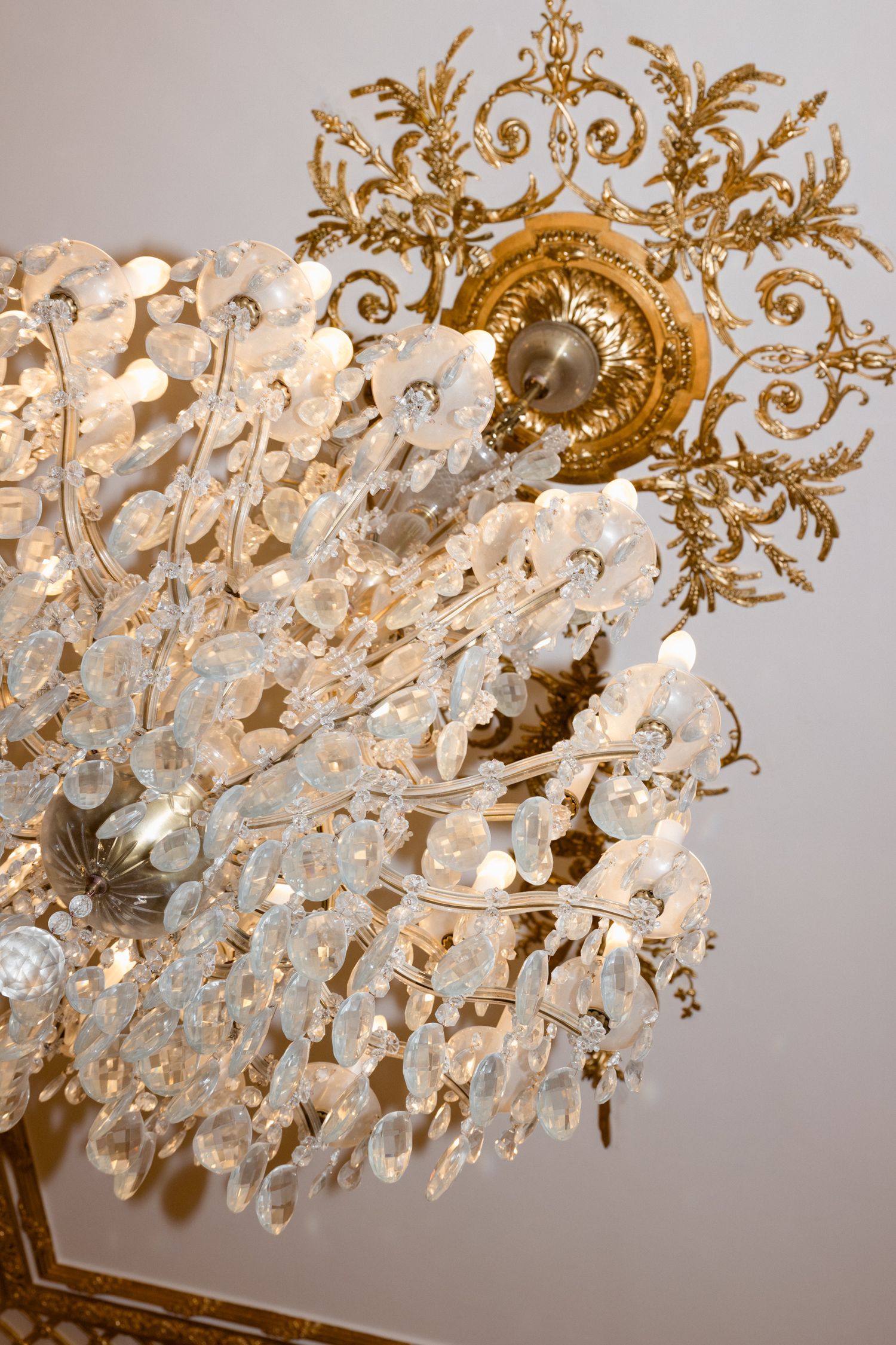 Ornate crystal chandelier with delicate glass flowers and gold filigree ceiling medallion captured in elegant detail. destination wedding fr