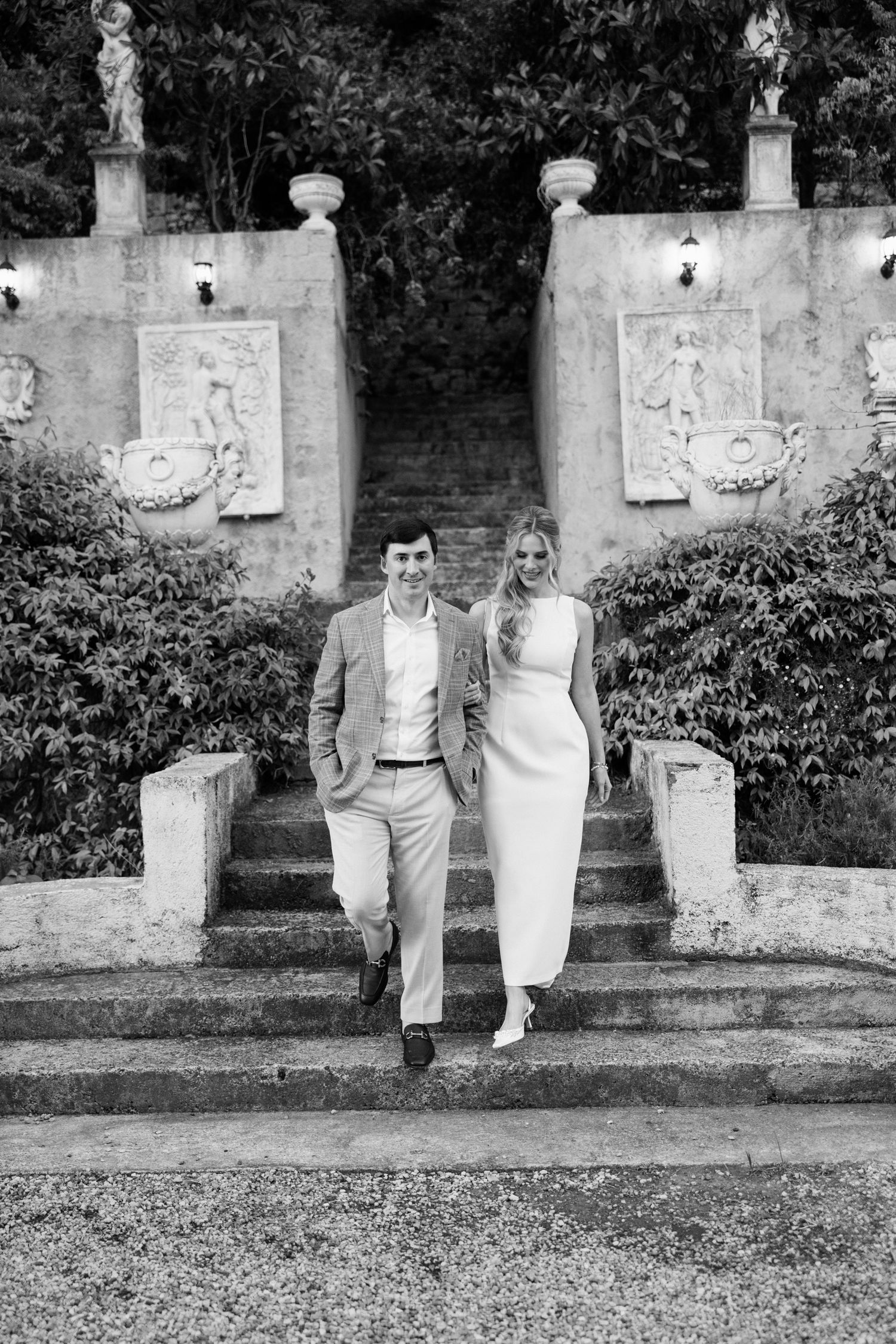 A couple stands together on stone garden steps with decorative wall panels and statues in a black and white photograph.