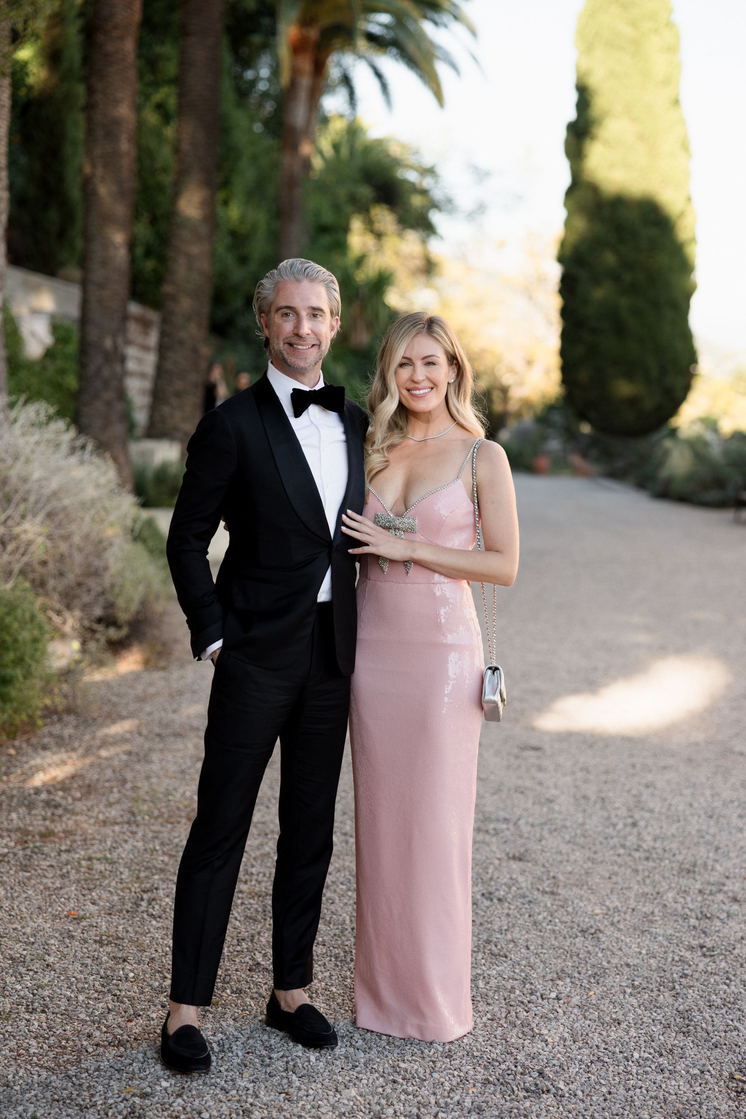 A couple in formal attire poses together on a palm tree-lined path during a sunset photoshoot.