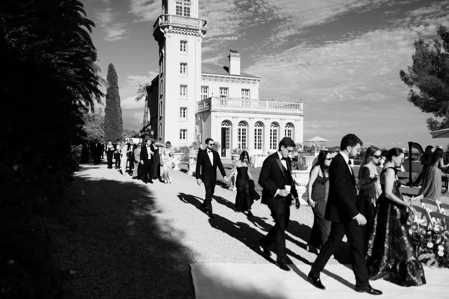 A group of formally dressed people walk in a line along a building exterior in black and white photography.