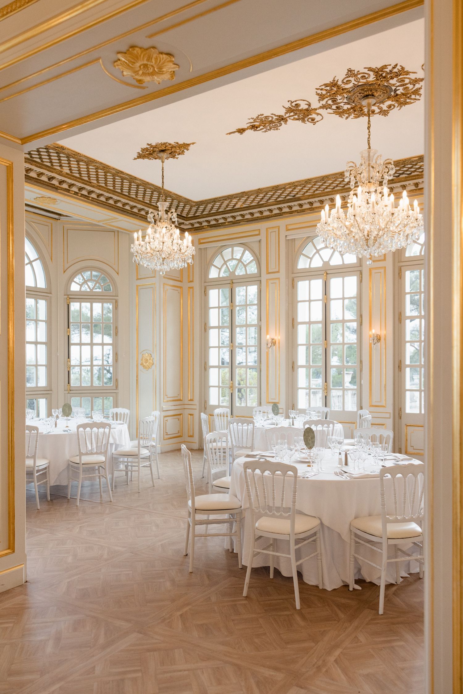 Elegant white and gold ballroom with crystal chandeliers, herringbone floors, and round dining tables set for a formal event.