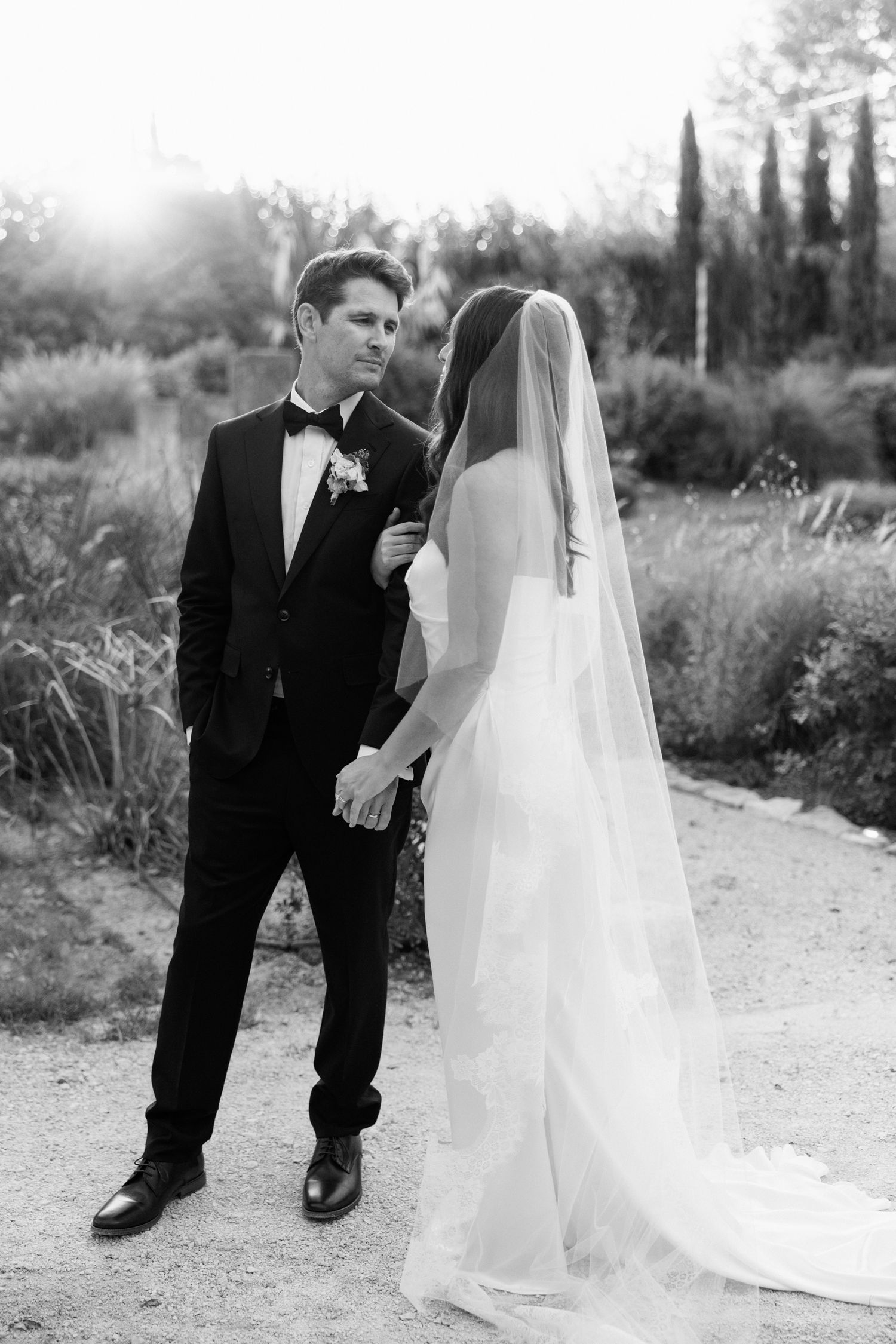 A black and white wedding photo of a couple standing outdoors with cypress trees and sunlight in the background.