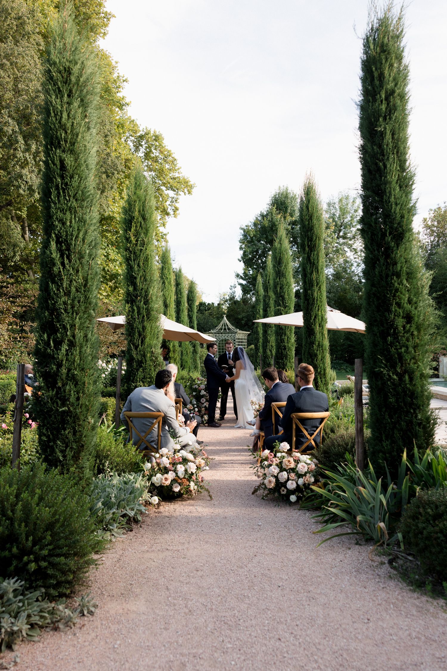 A scenic outdoor path lined with tall cypress trees and white umbrellas creates an elegant garden walkway setting. wedding photographer