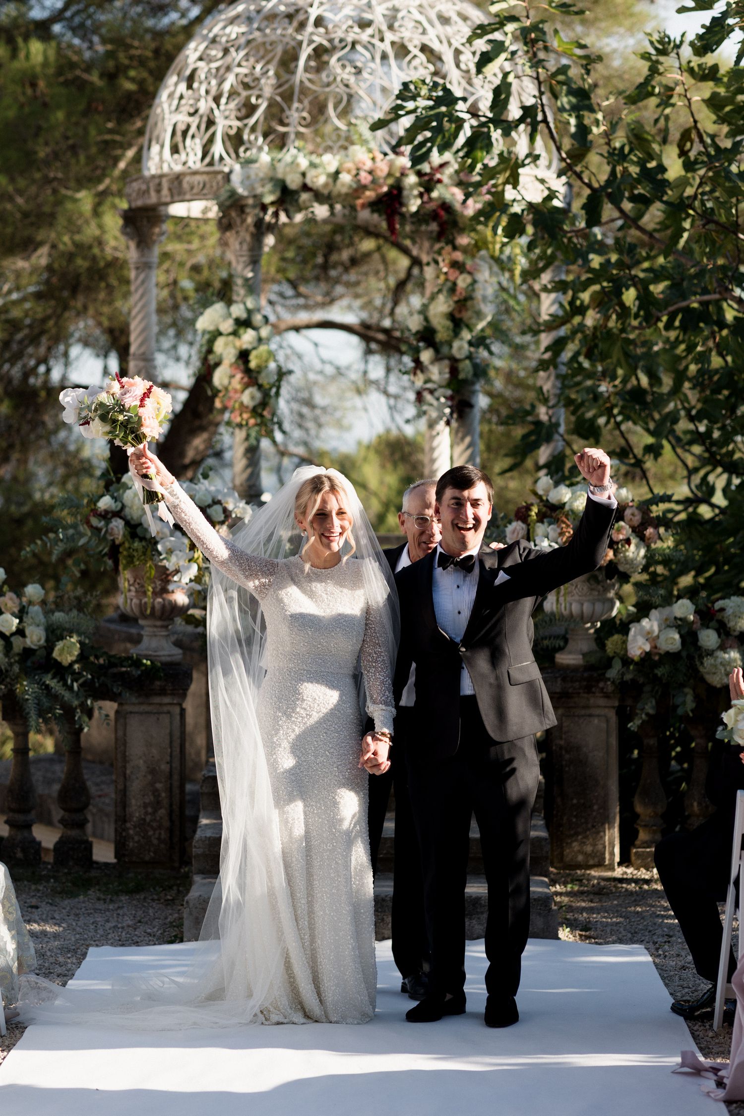 Couple celebrates joyfully under a white floral arch during an elegant outdoor garden wedding ceremony.