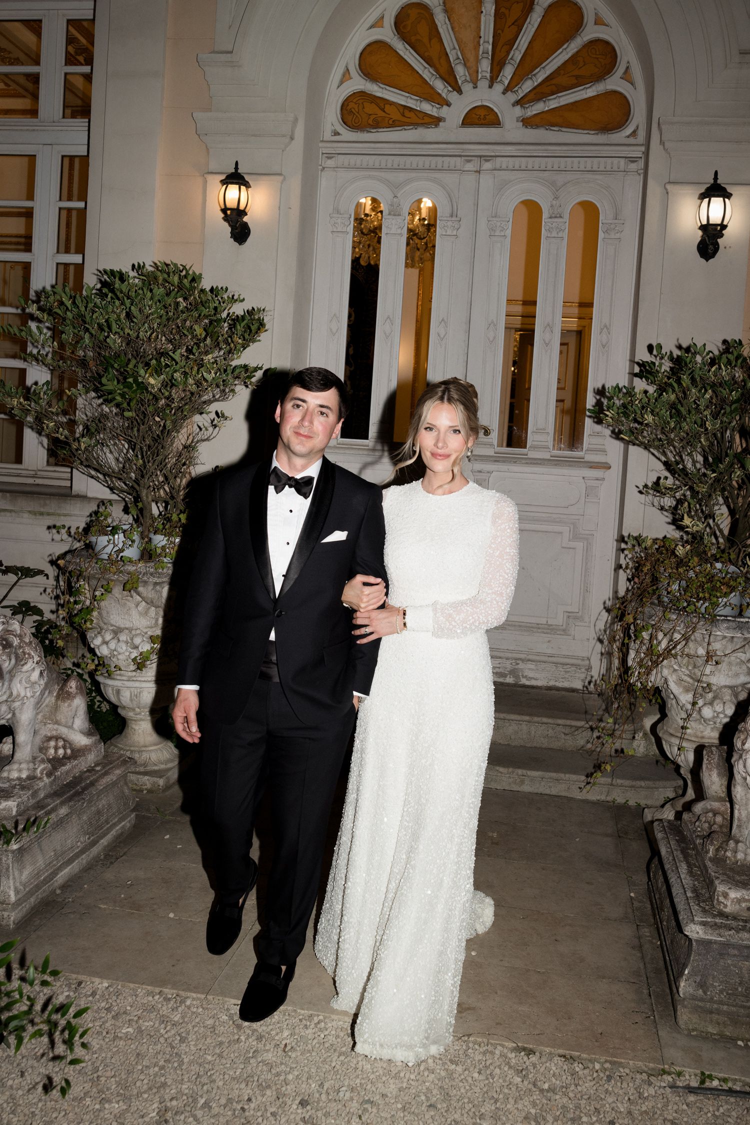 A couple in formal wedding attire walks arm in arm outside an elegant white building with a decorative sunburst design.