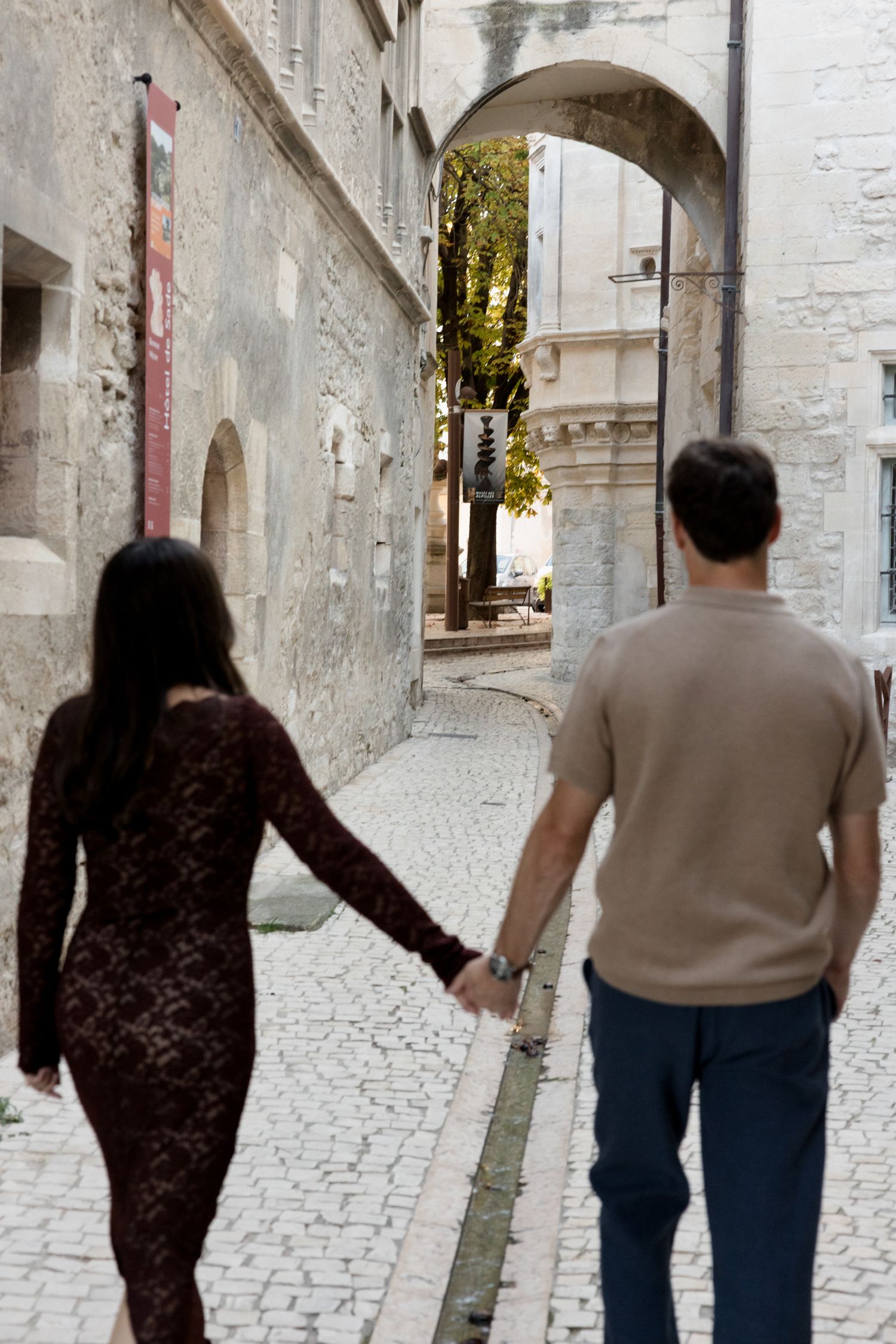 Two people walk down a narrow cobblestone alley with white stone walls and an arched passageway ahead.