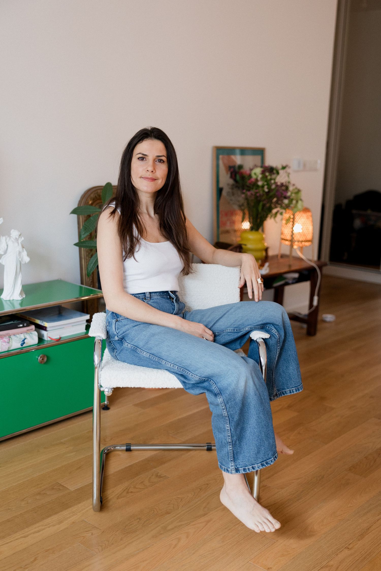 Woman in white tank top and jeans sitting barefoot on a chair in a cozy, well-lit living room.