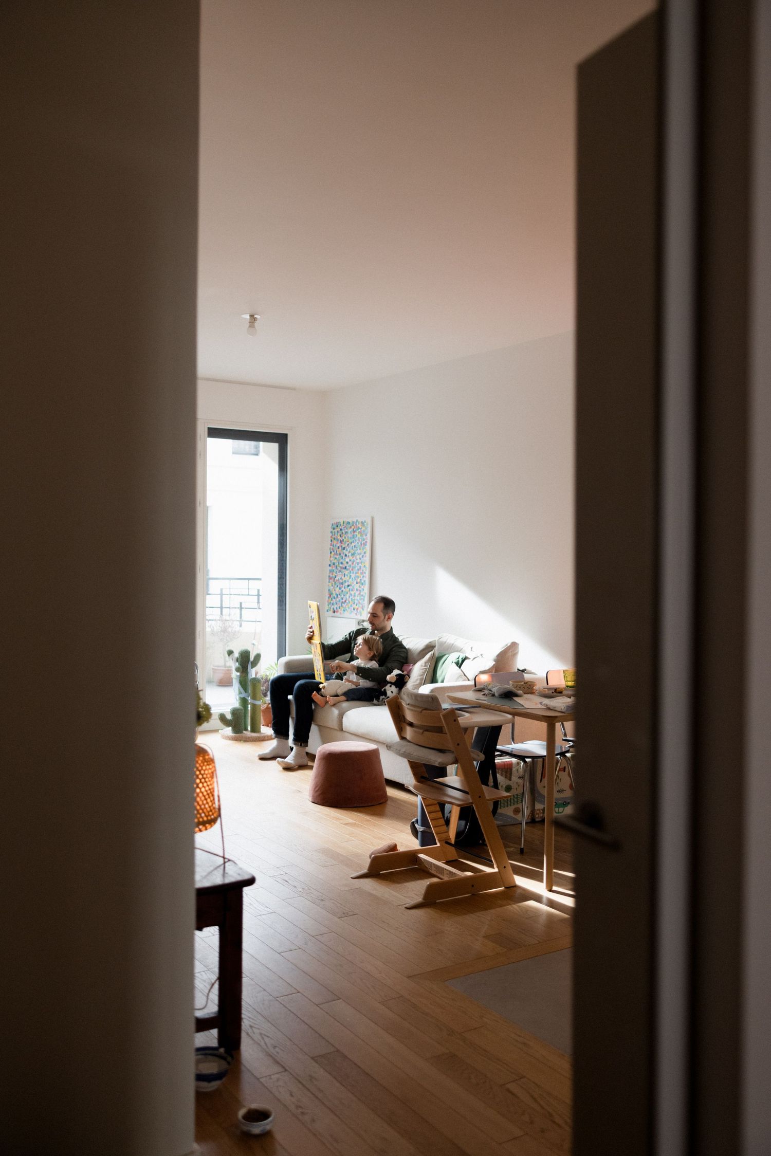 Person relaxing on a sofa in a bright, modern living room with wooden floors, viewed through an open doorway.