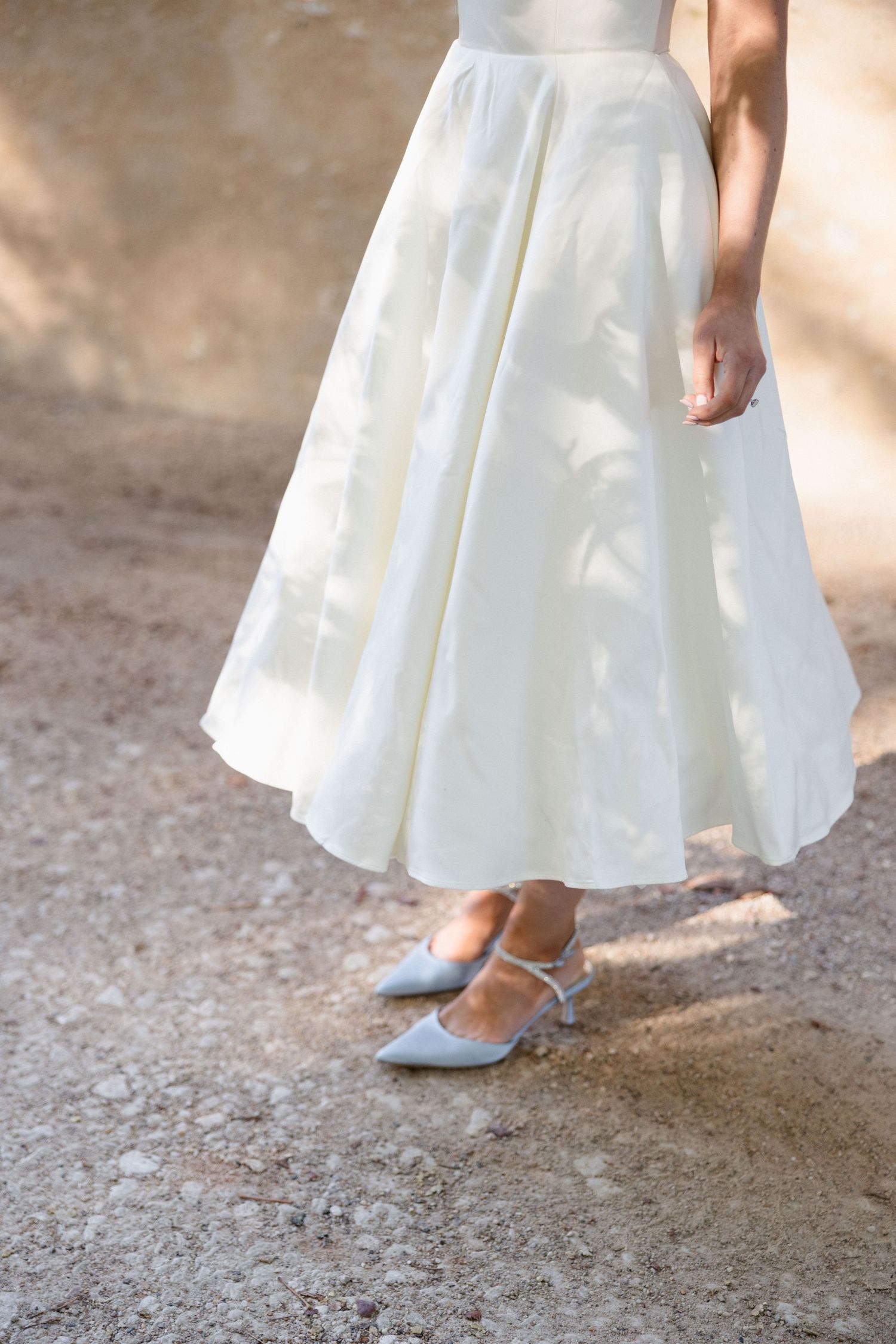 Close-up of bride in white midi wedding dress and silver kitten heel shoes on gravel path.