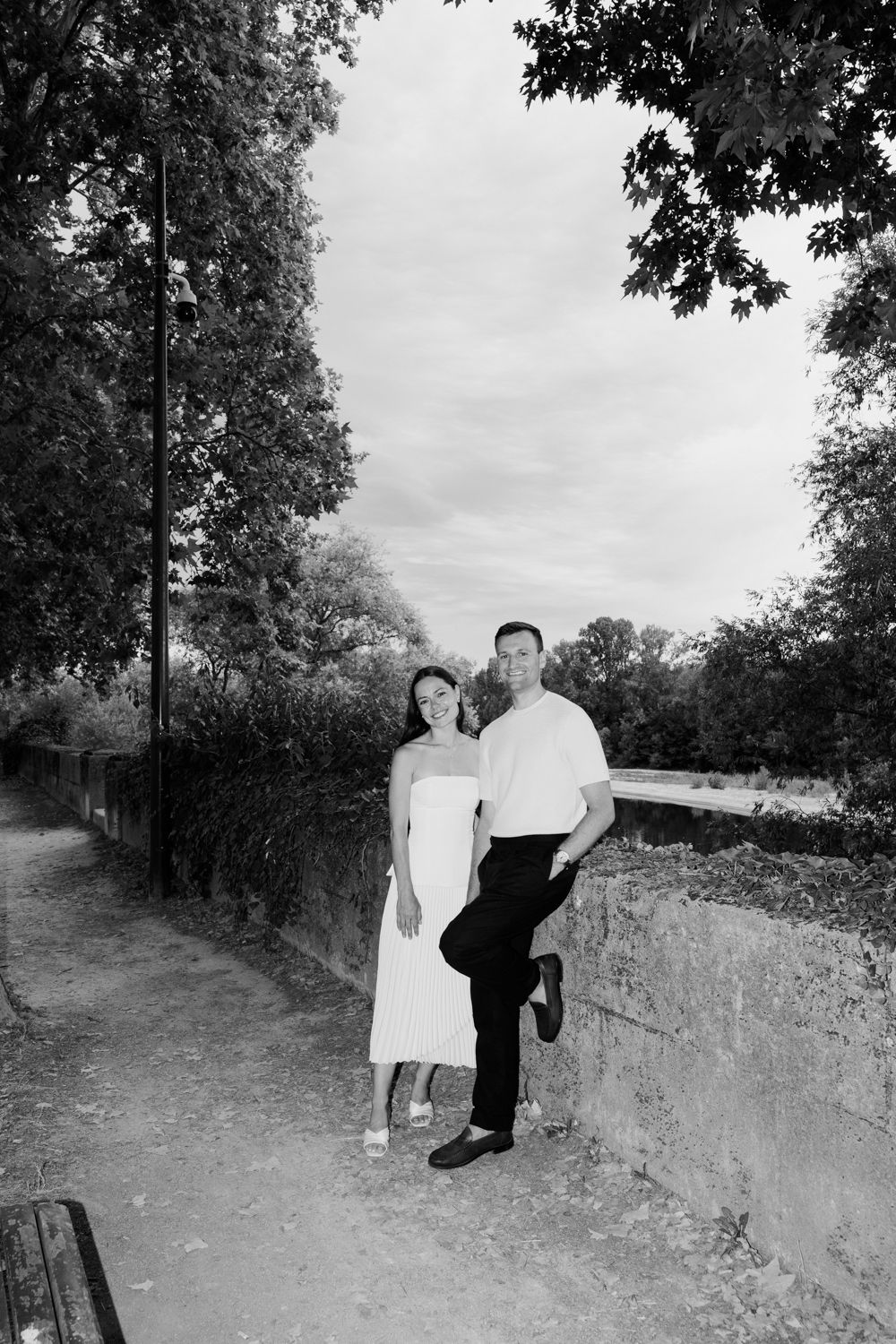 A vintage black and white photograph shows a couple standing together on a tree-lined path beside a stone wall. Engagement session in Tours