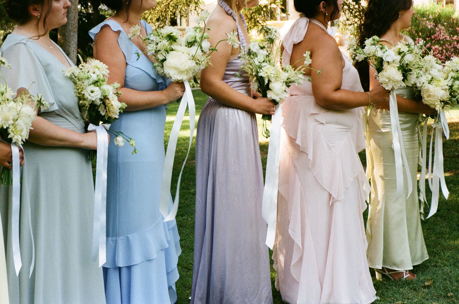 A row of bridesmaids in pastel blue and blush dresses hold white floral bouquets at an outdoor wedding ceremony. Wedding on film, analog wed