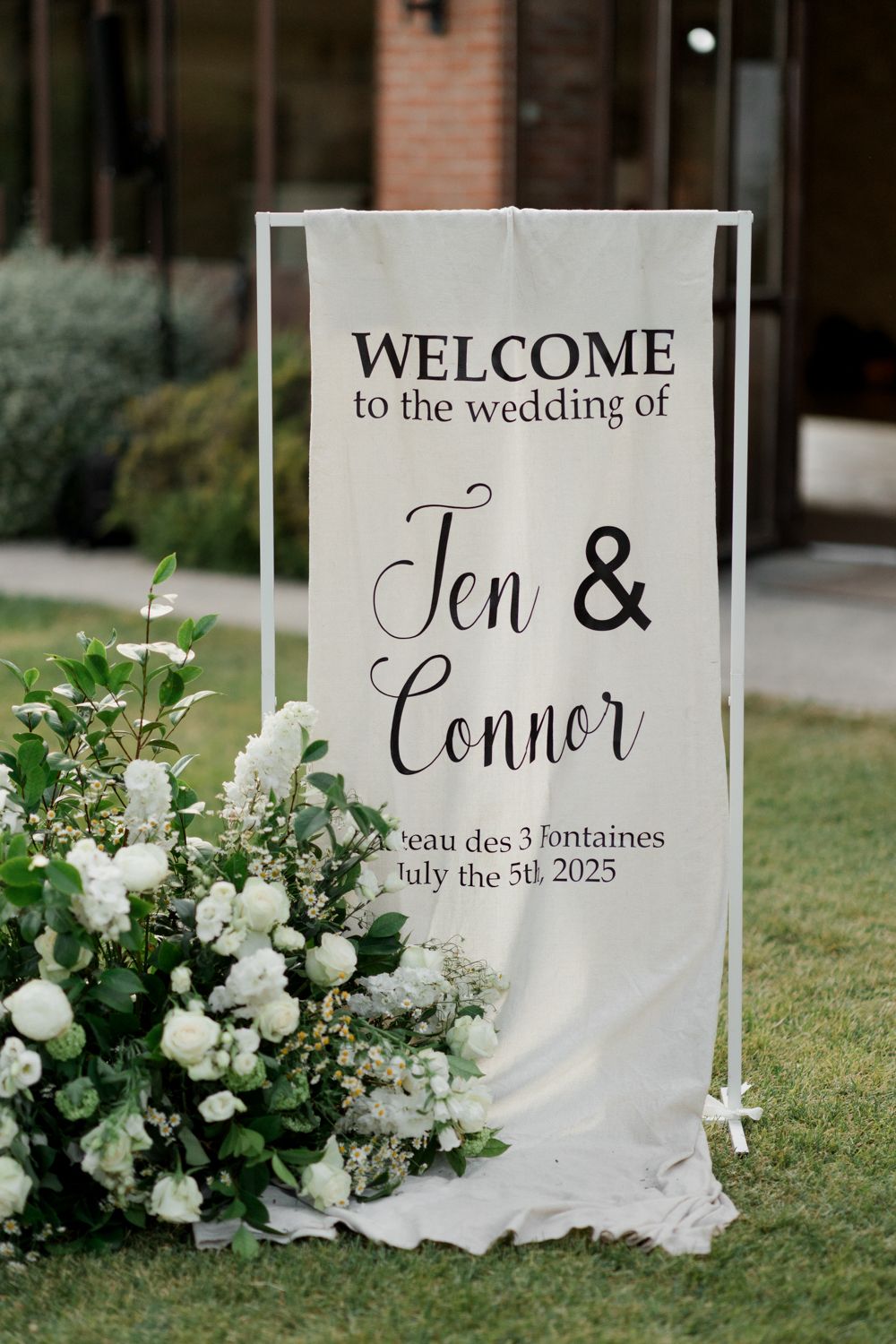 Elegant white wedding welcome signs with calligraphy text and floral arrangements in an outdoor garden setting. Château des 3 Fontaines wedd