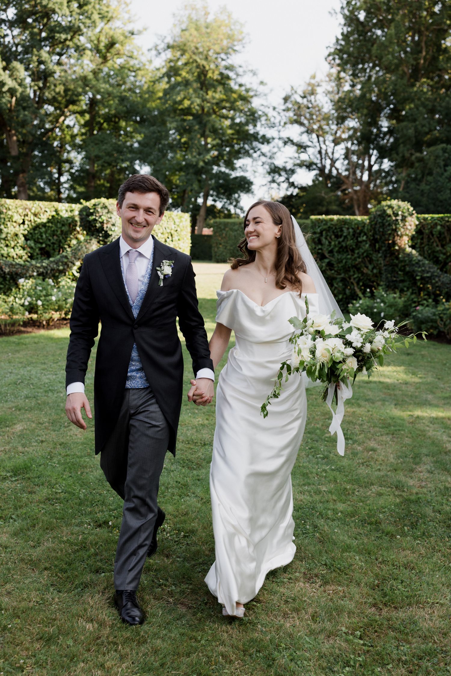 A newlywed couple walks and laughs together on a grassy lawn during their outdoor wedding photoshoot.