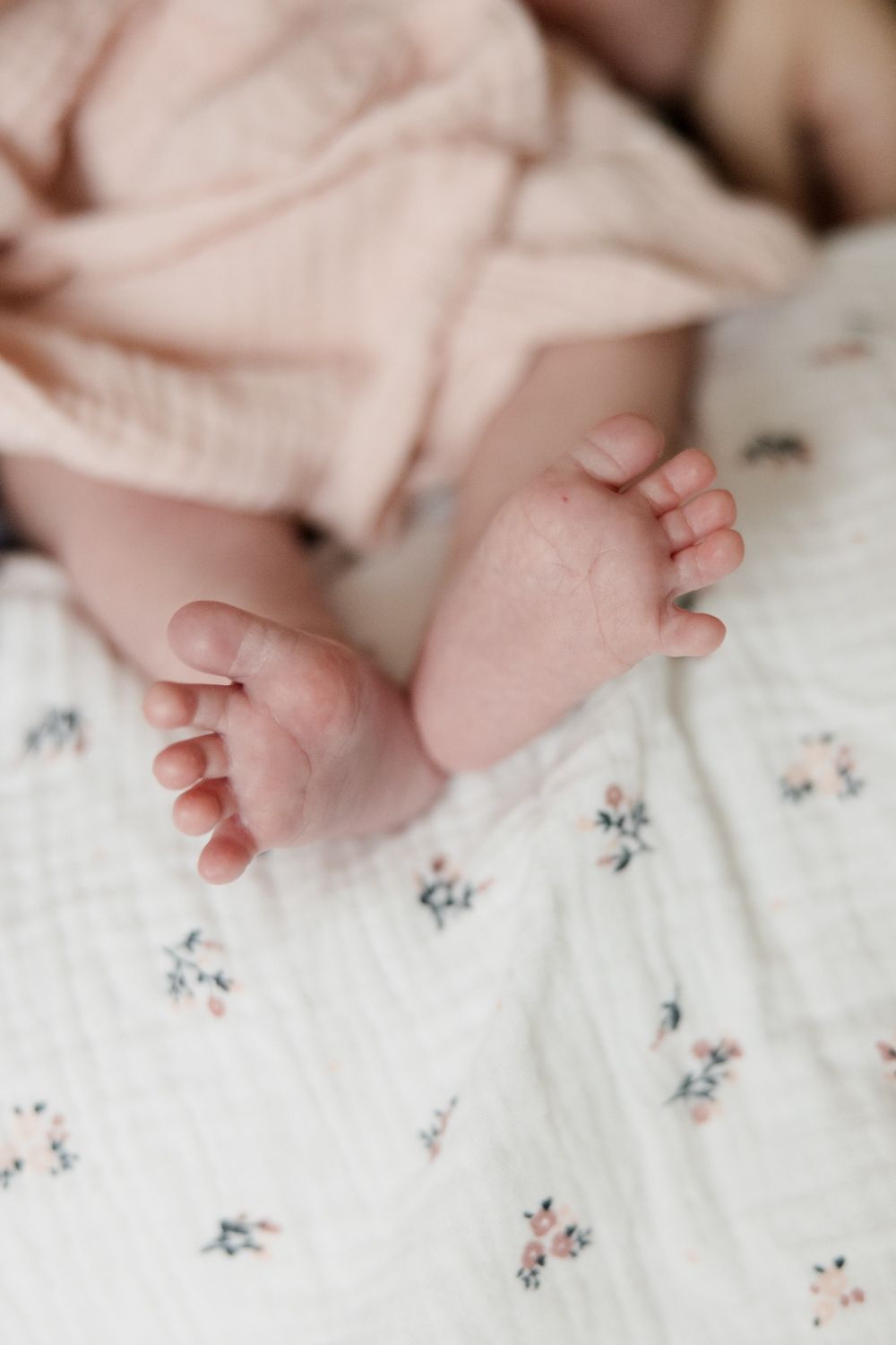 Tiny newborn feet rest on white floral patterned bedding with a soft pink blanket in frame.