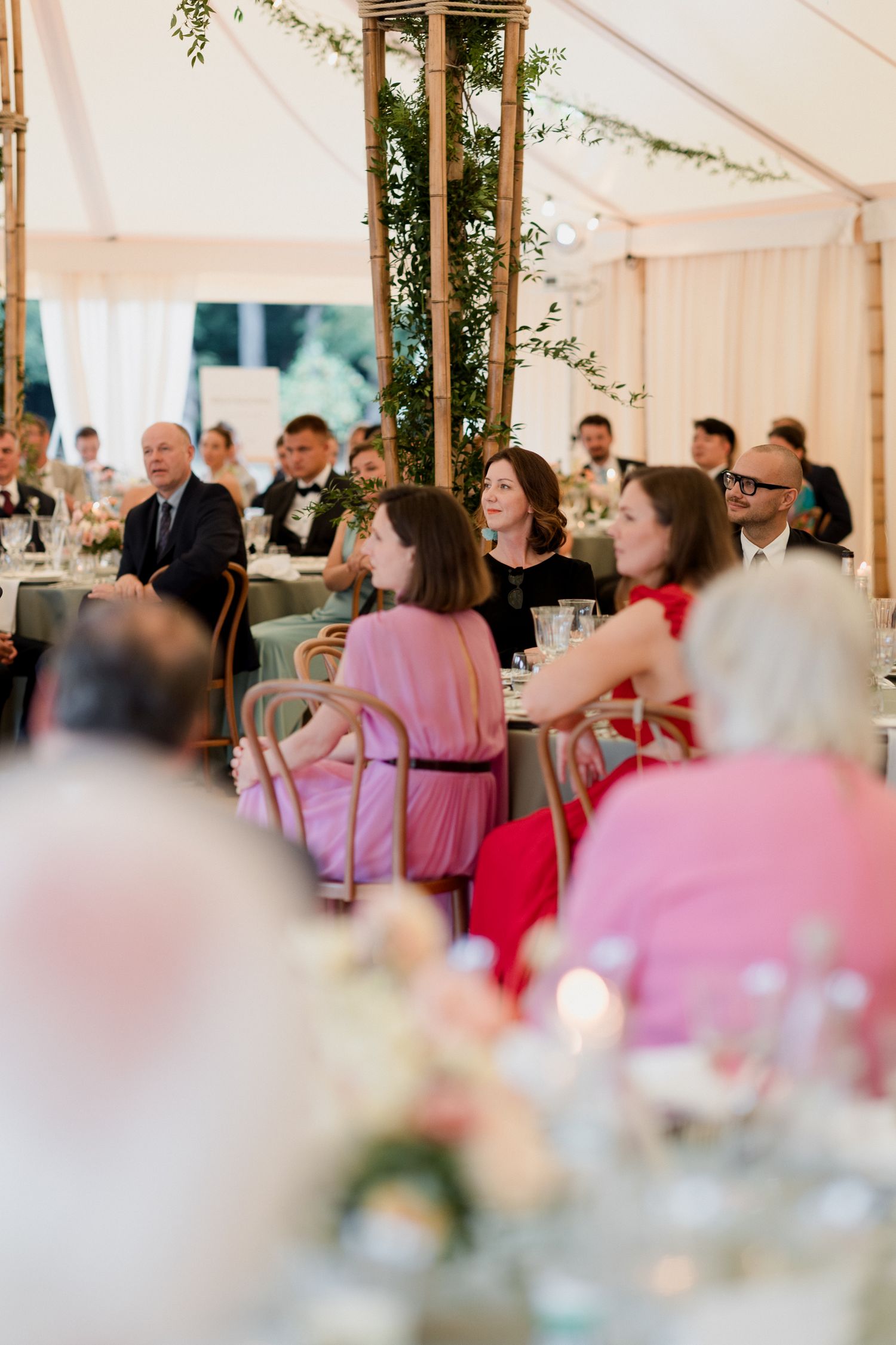 Wedding guests seated at elegant tables under a white tent with floral decorations and pink accents.
