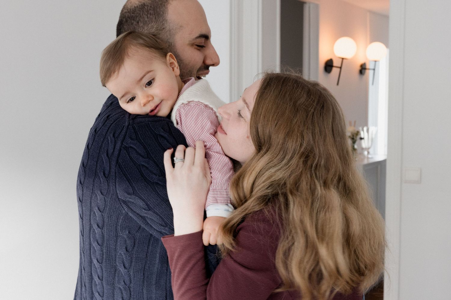 A happy family with a father holding a baby while a mother kisses the infant in a bright home interior.