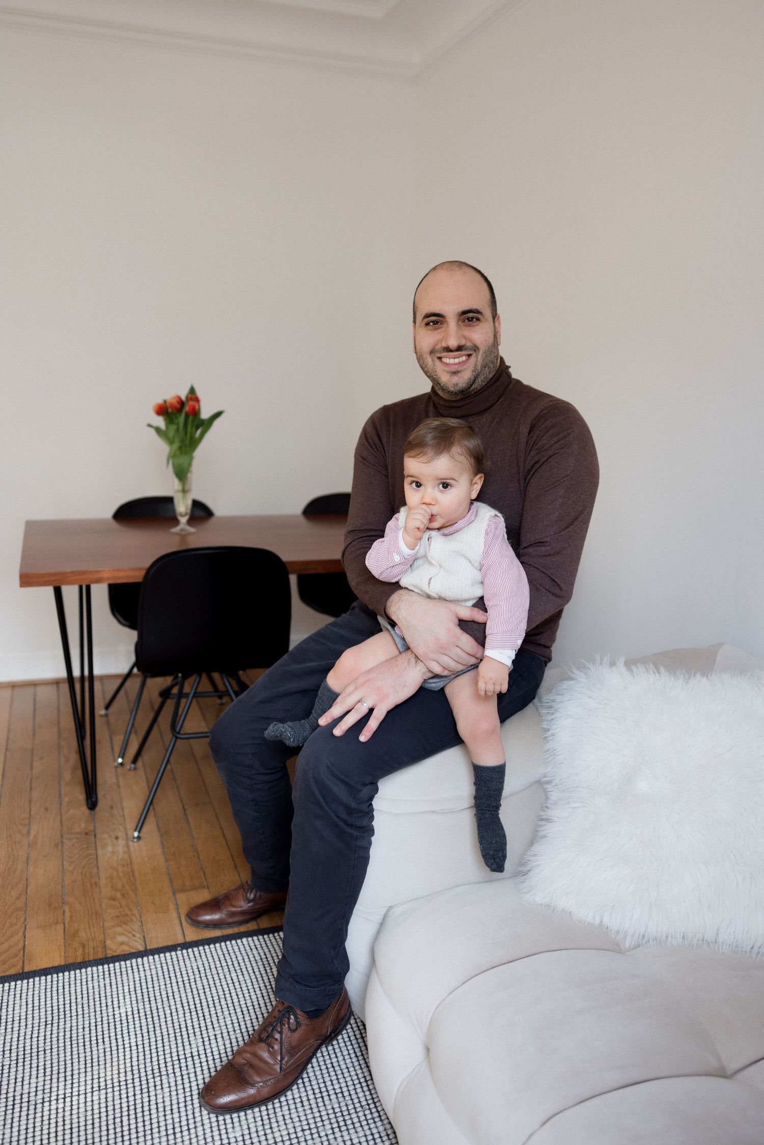 A smiling man in a brown turtleneck sits on a white sofa holding a baby in a bright modern living room.