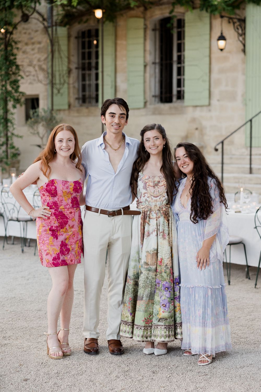 Family group poses together outdoors in coordinated outfits on a gravel path by a stone villa with flowing dresses and florals.
