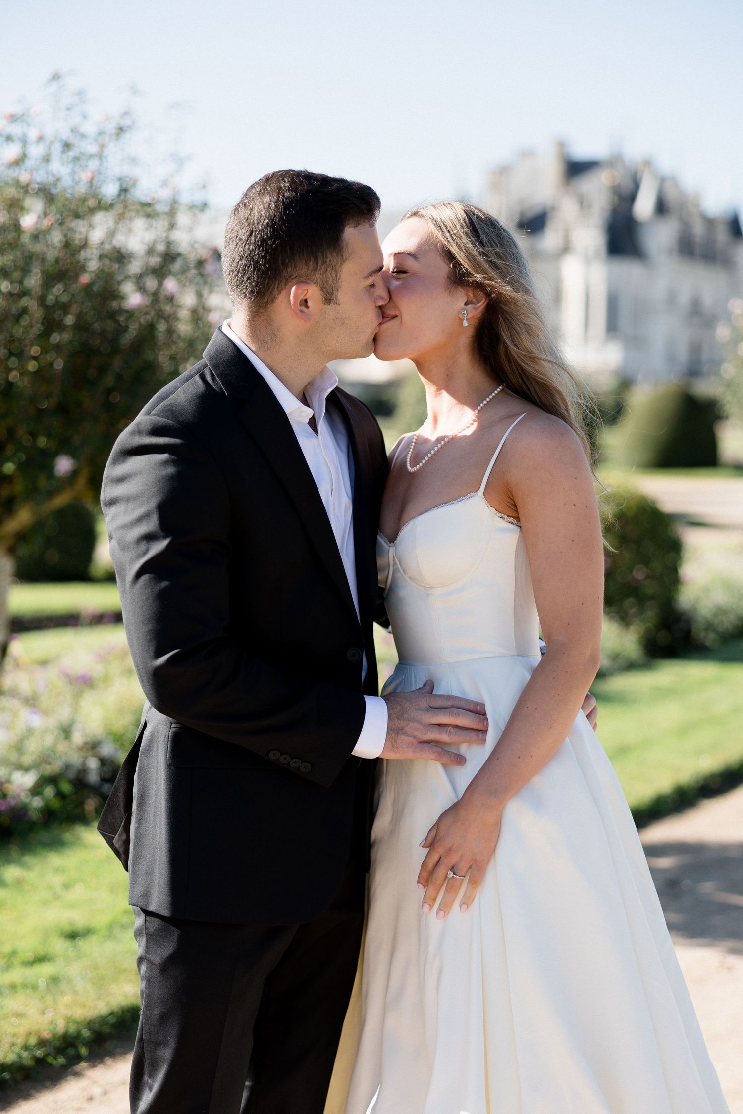 A bride and groom share a romantic kiss in a sunlit garden with a grand chateau in the background.