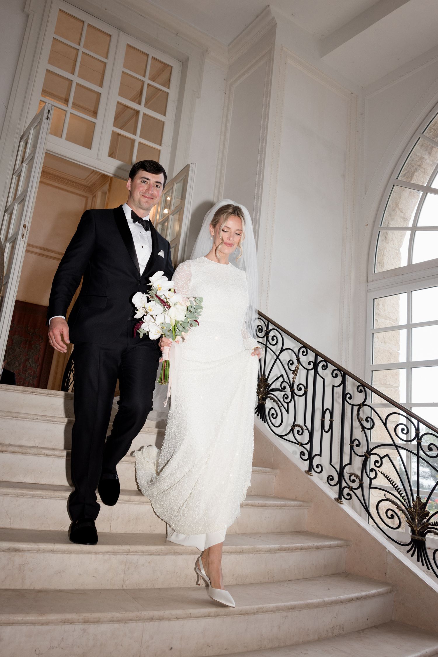 A couple descends an elegant white marble staircase with ornate iron railings on their wedding day.