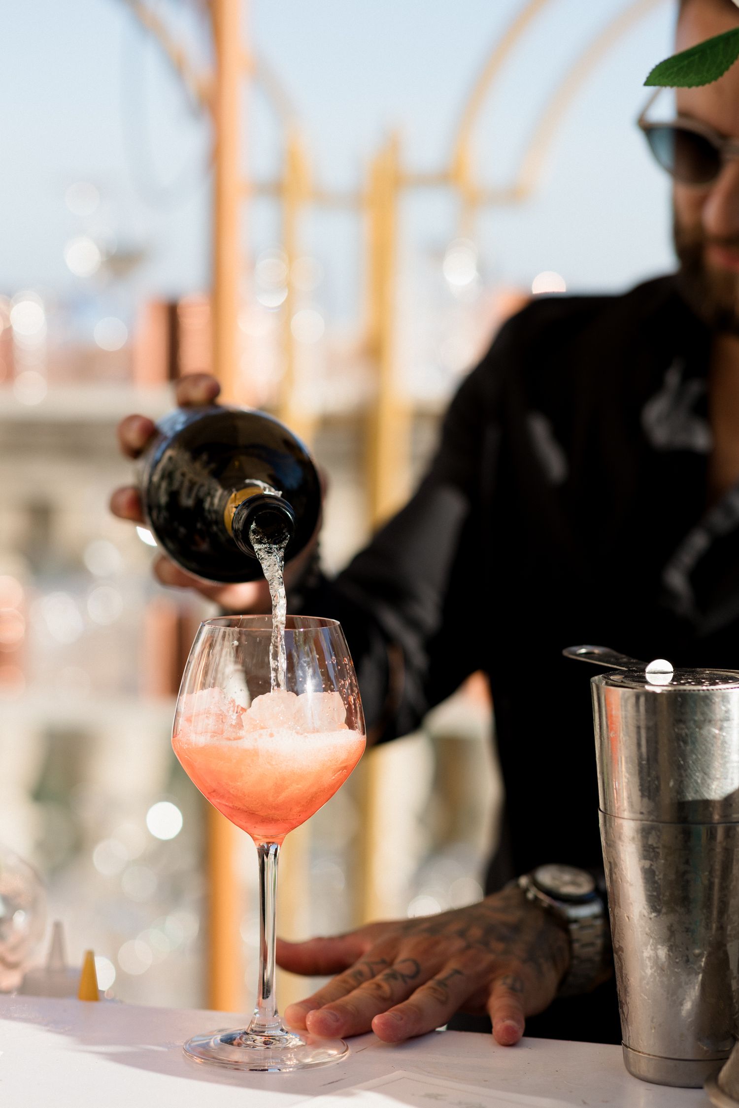 A bartender pours a pink cocktail into a wine glass next to a silver cocktail shaker on a white bar counter.