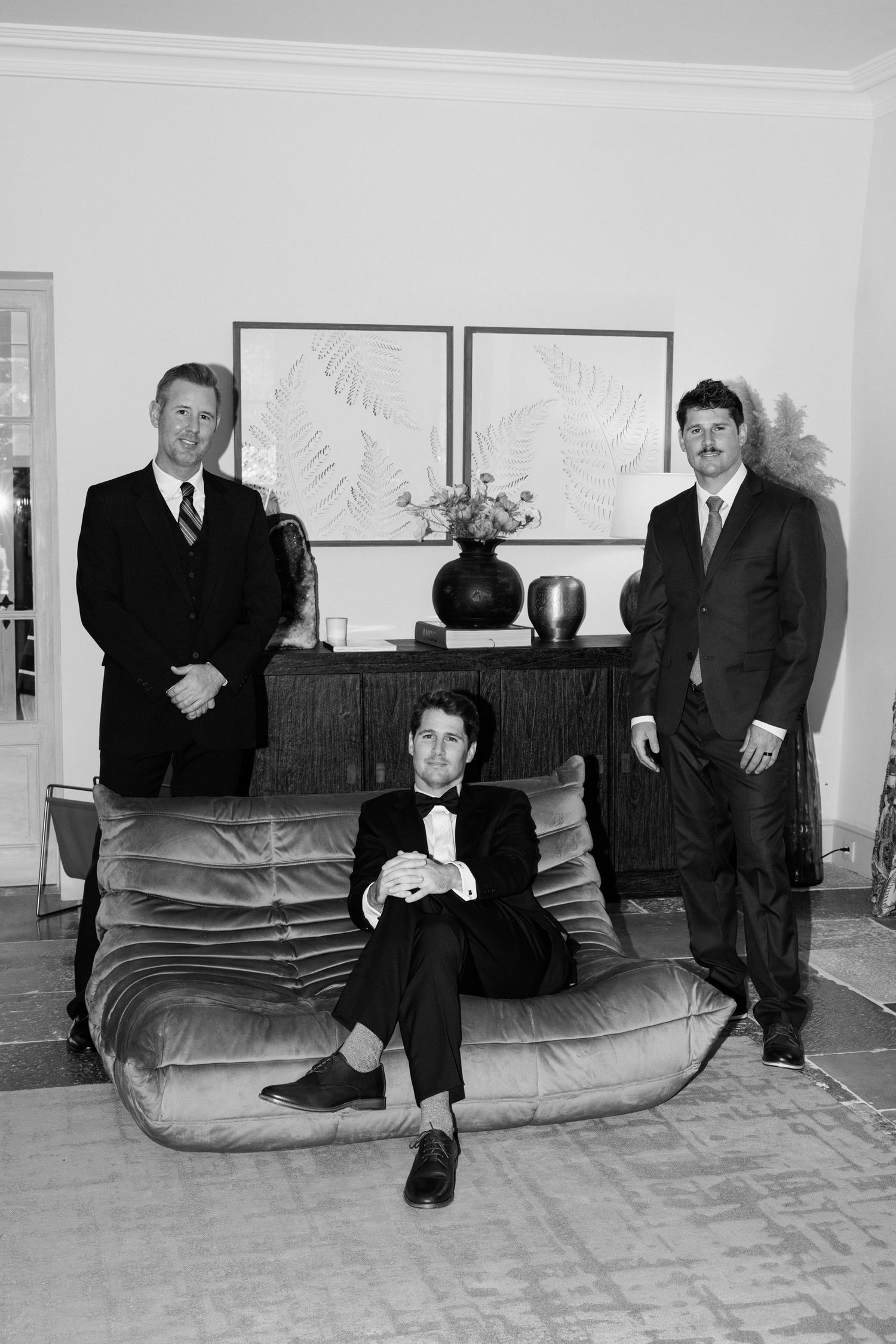 Three men in suits pose in a living room with a cushioned sofa and framed artwork on the wall in black and white. Editorial wedding photogra