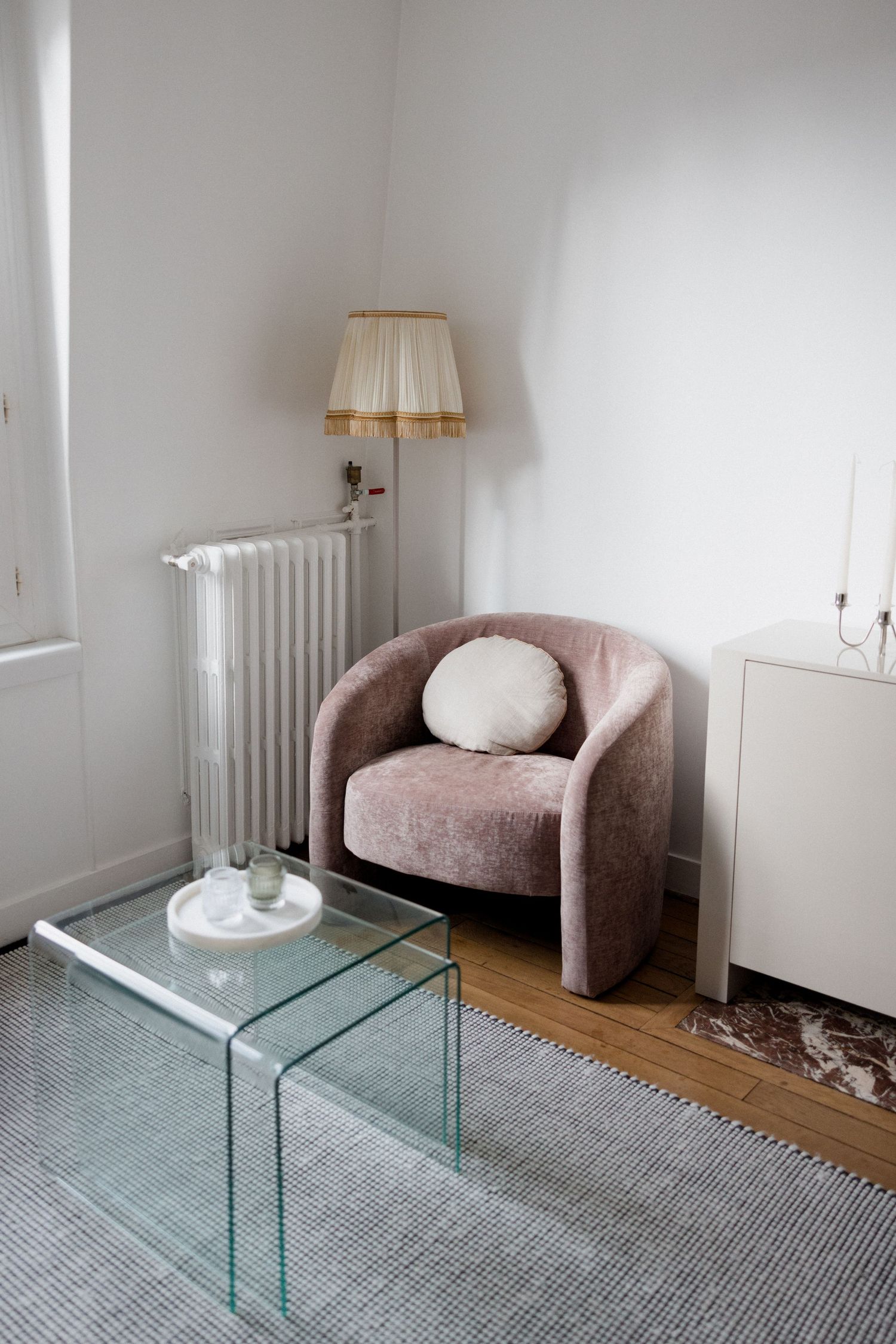 Minimalist living room corner with a dusty pink velvet armchair, glass nesting tables, floor lamp, and white radiator.