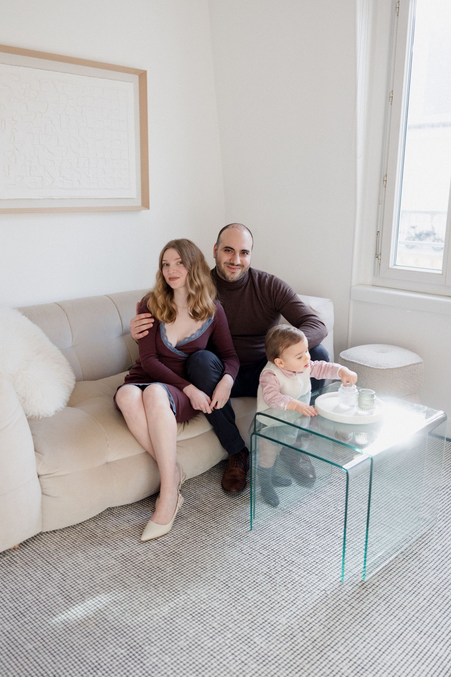 A happy couple sits on a beige sofa with their toddler playing at a glass coffee table in a bright living room.