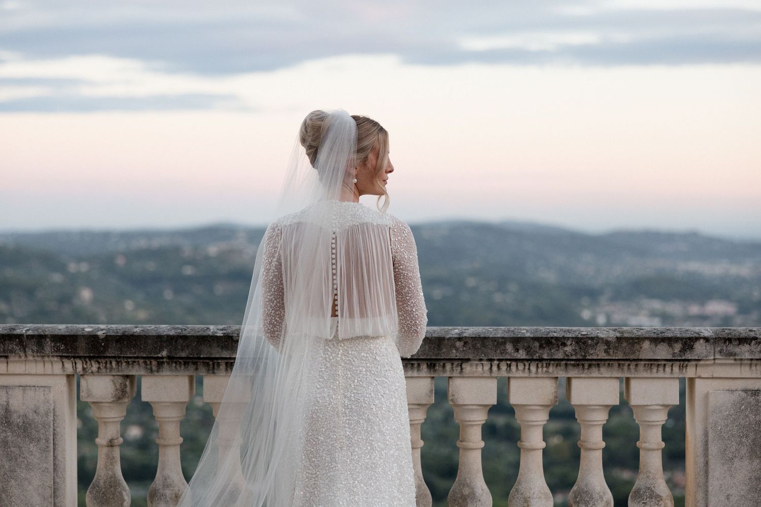 A bride in a flowing veil stands on a stone balcony overlooking scenic mountain views at sunset. Mariage Chateau saint georges Grasse France