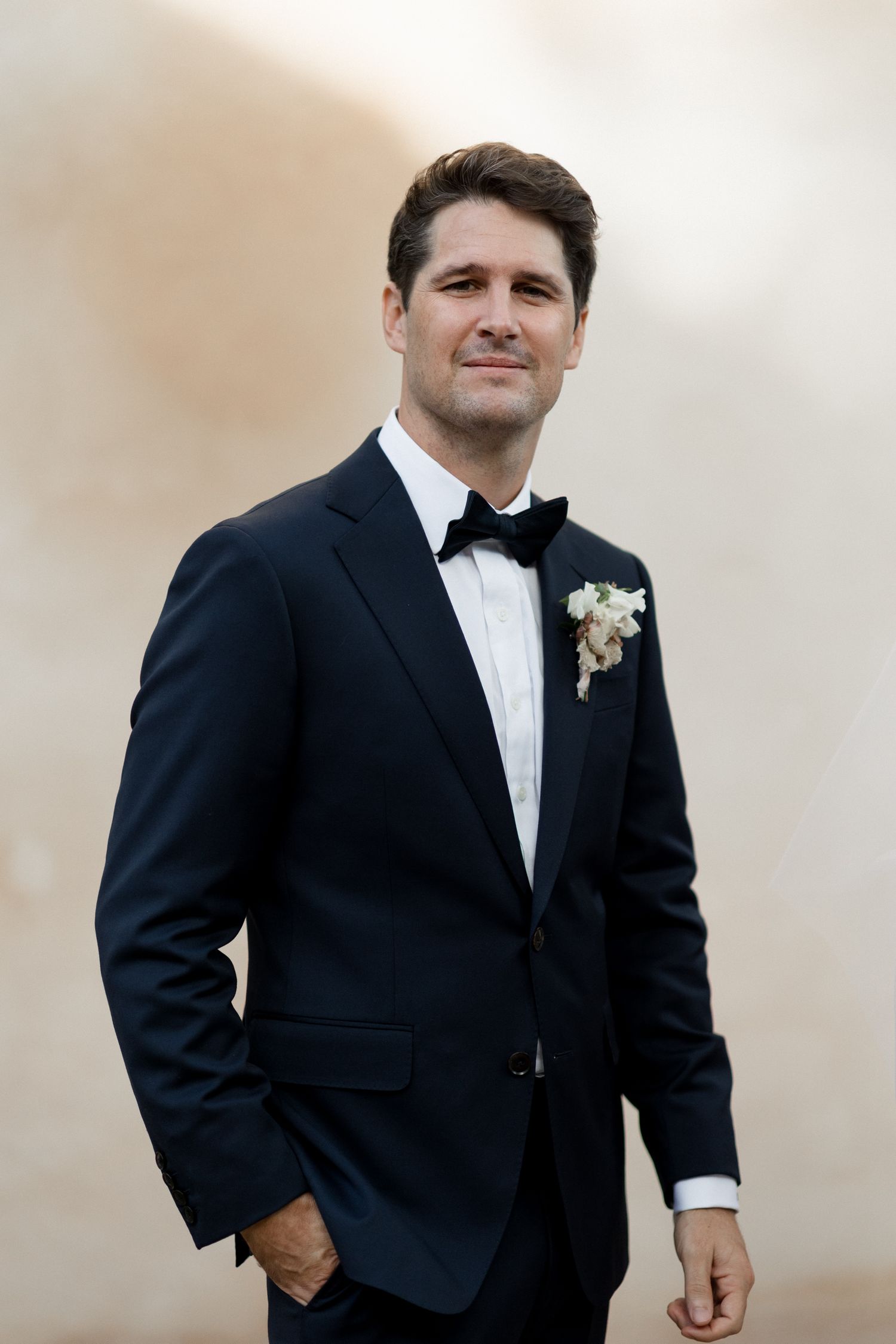 Groom in classic black tuxedo and bow tie poses for formal wedding portraits against neutral background.