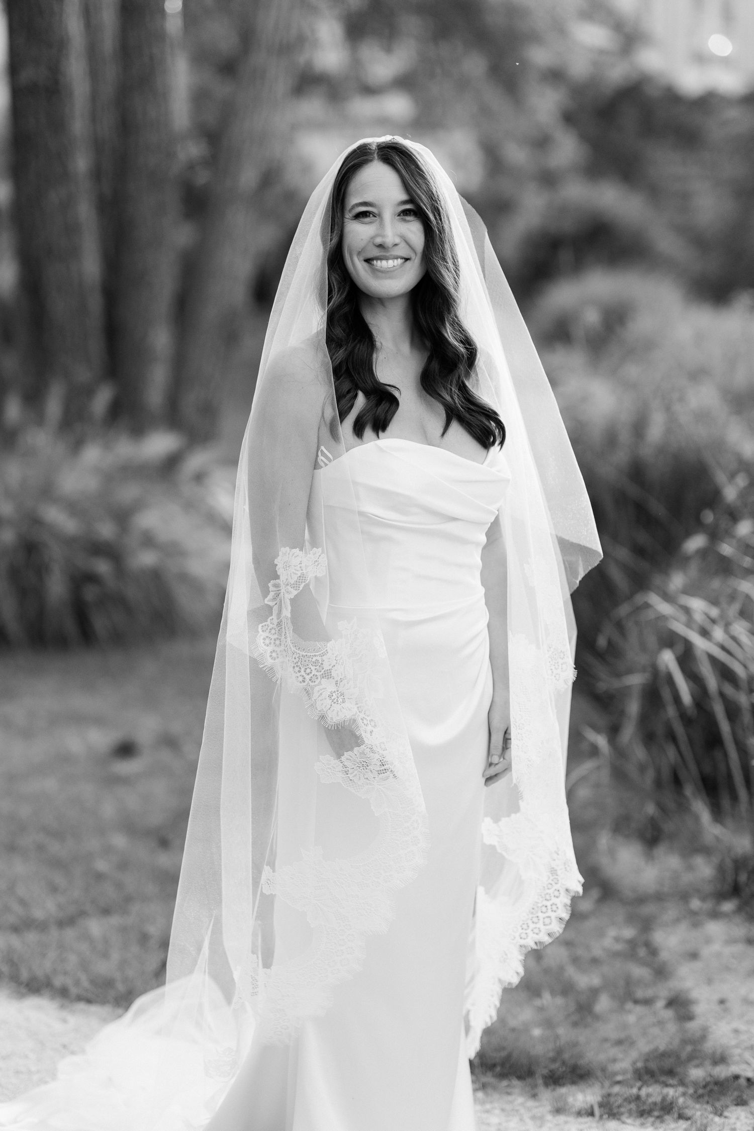 A bride in a flowing white wedding dress and veil poses outdoors in a garden setting, creating a romantic bridal portrait.