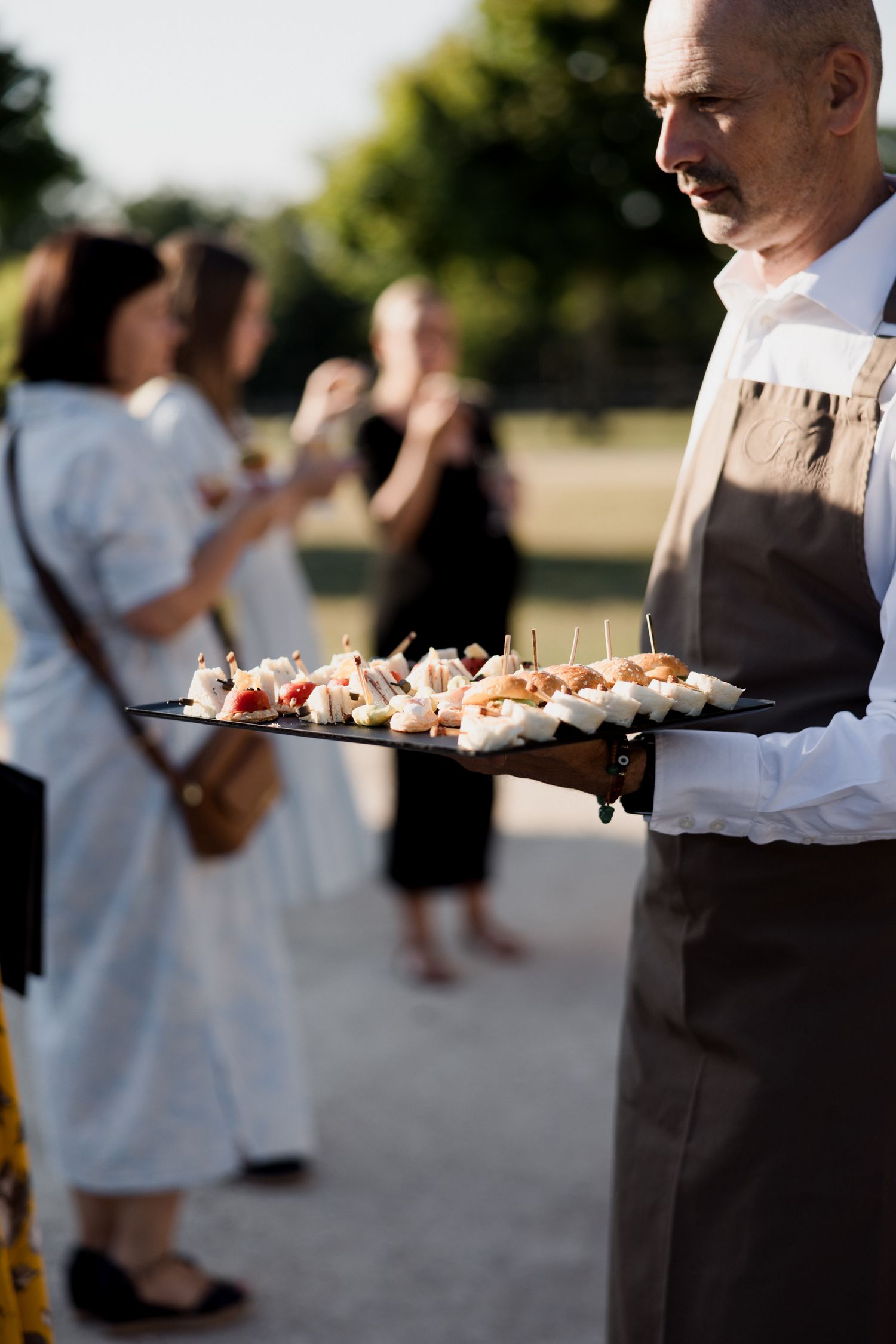 Servers carry appetizer trays at an outdoor catering event during golden hour.