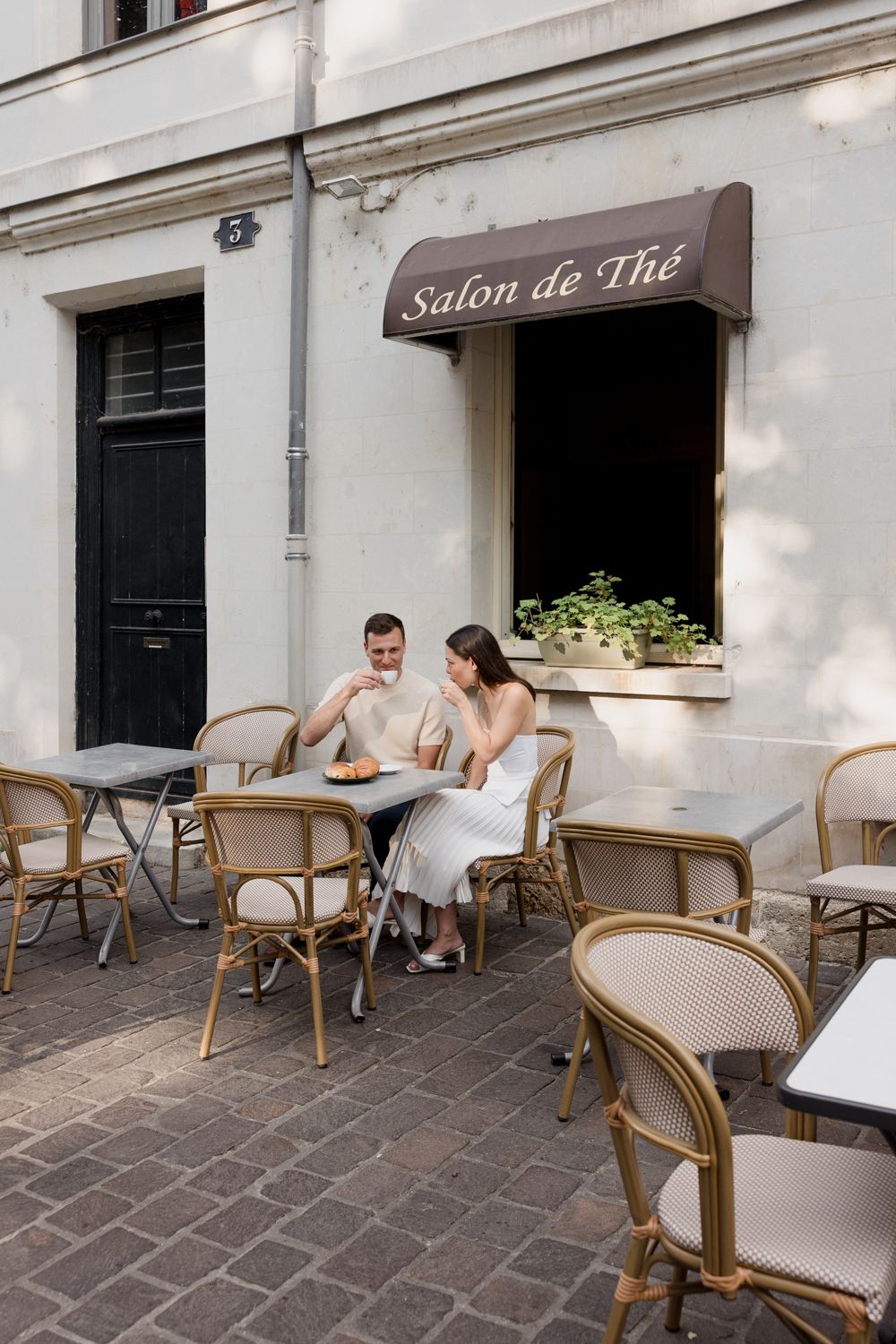 Cafe patrons sit at rattan chairs and tables outside a quaint Salon de The with white walls and cobblestone paving.