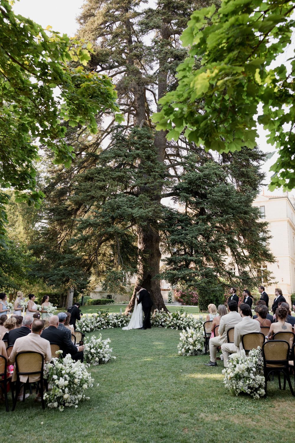 An outdoor wedding ceremony in progress on a lush green lawn with tall trees and white floral arrangements.