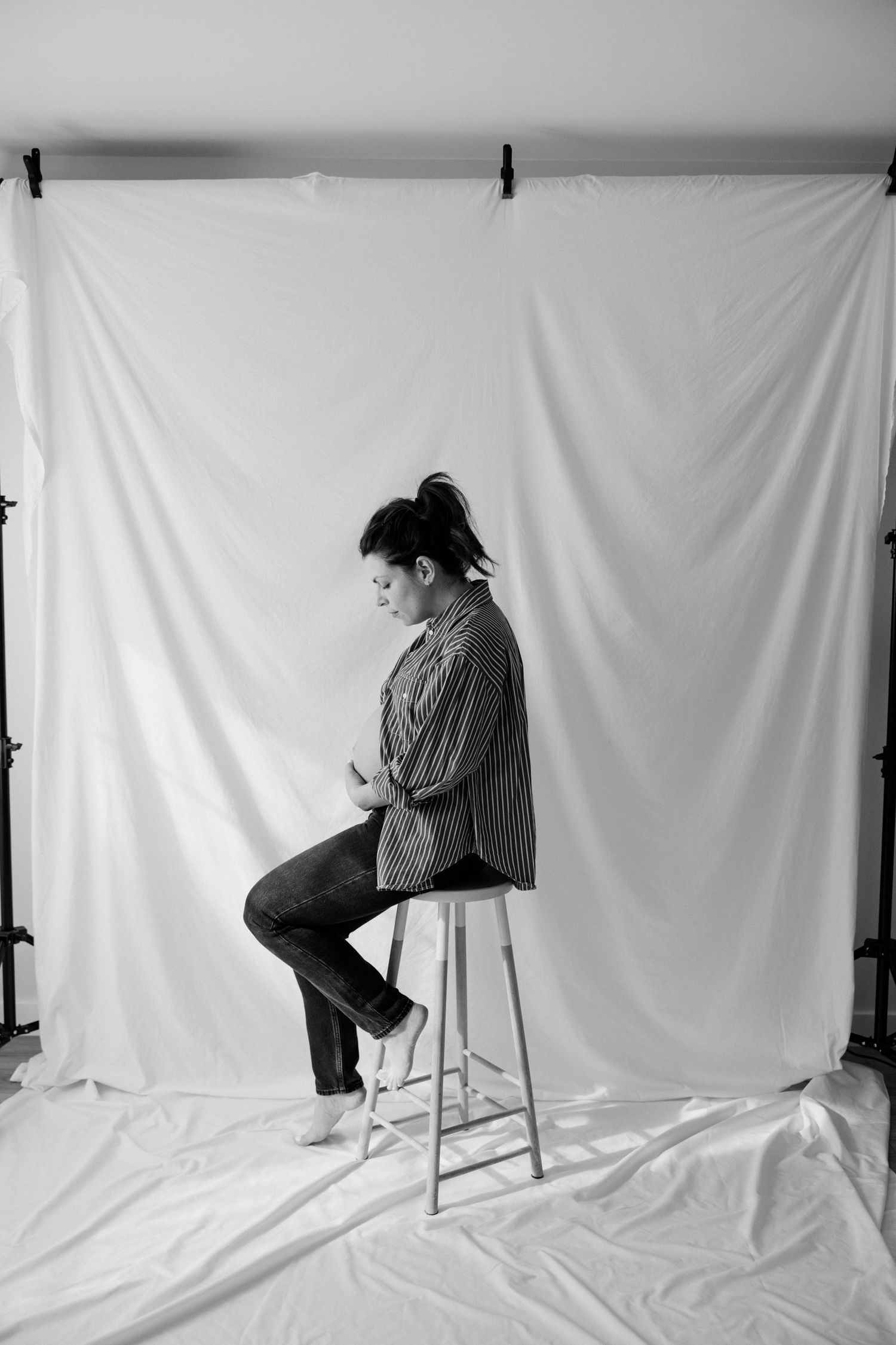Black and white portrait photograph of someone seated on a stool against a white backdrop wearing casual clothing.