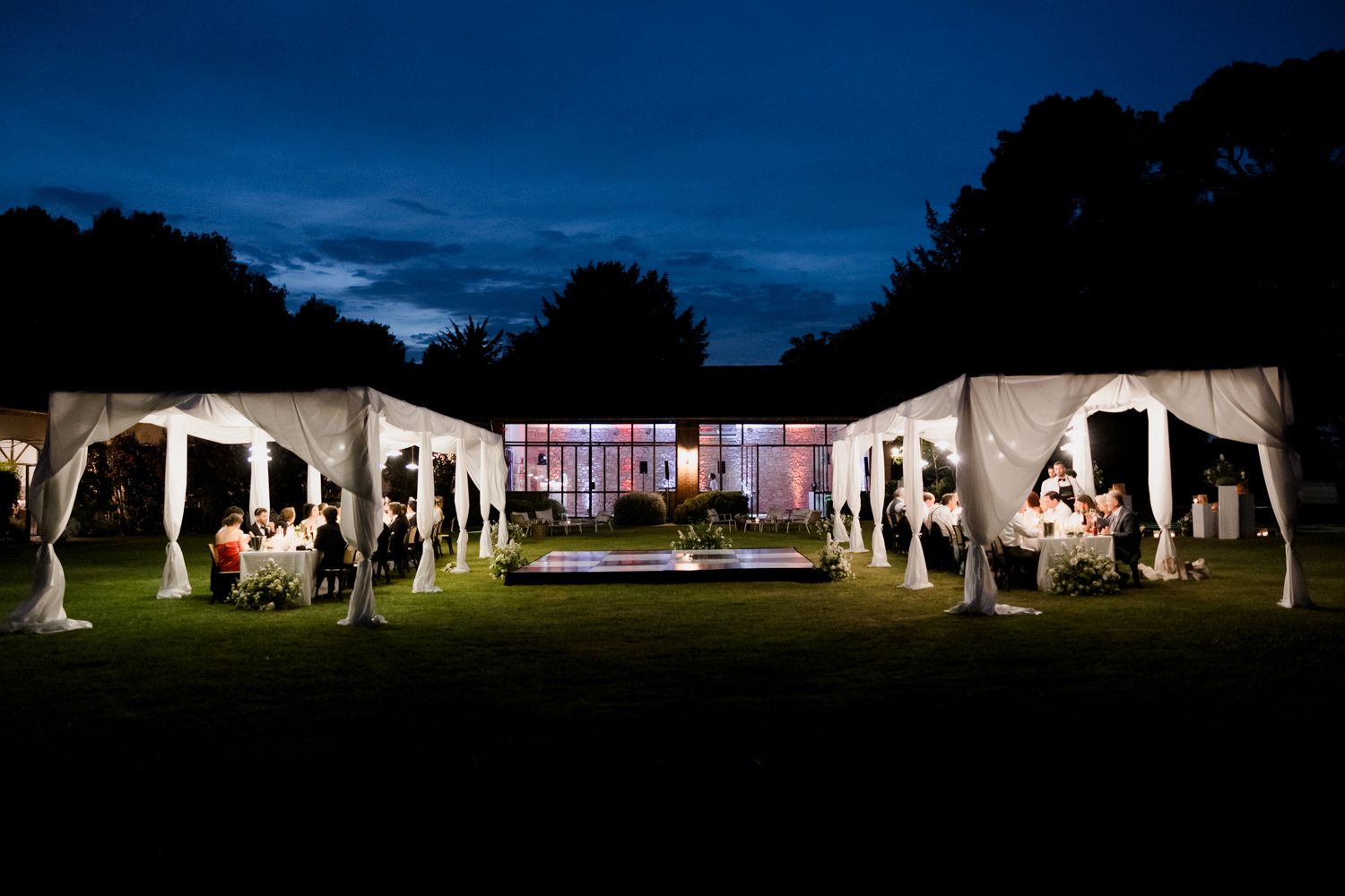 Long exposure nighttime panorama of an outdoor wedding reception tent with glowing lights and guests.