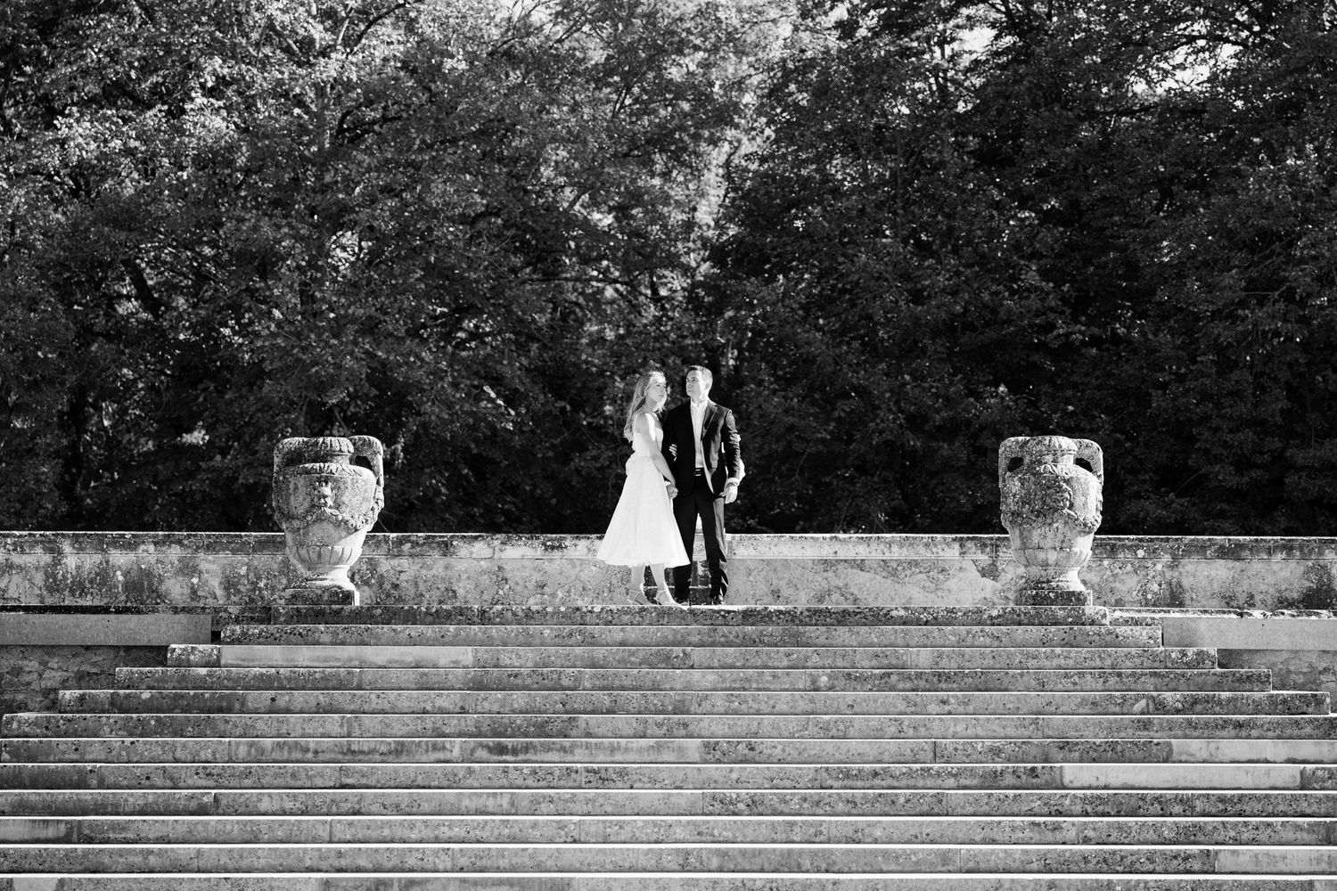 Black and white photo of a bride and groom standing atop grand stone steps surrounded by lush trees and ornate urns.