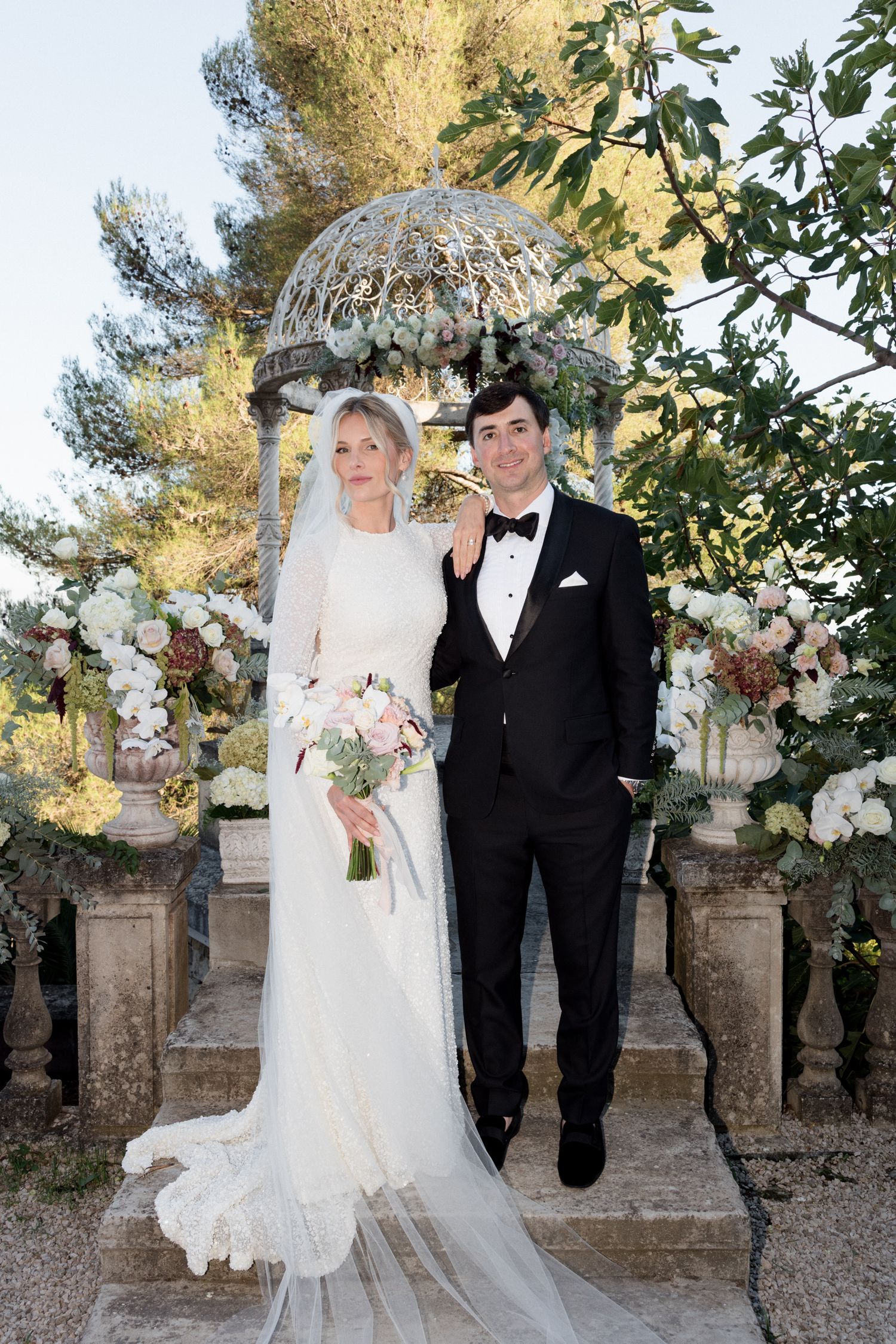 A couple poses together in formal wedding attire against a stone wall backdrop with lush floral arrangements. Photographe mariage france