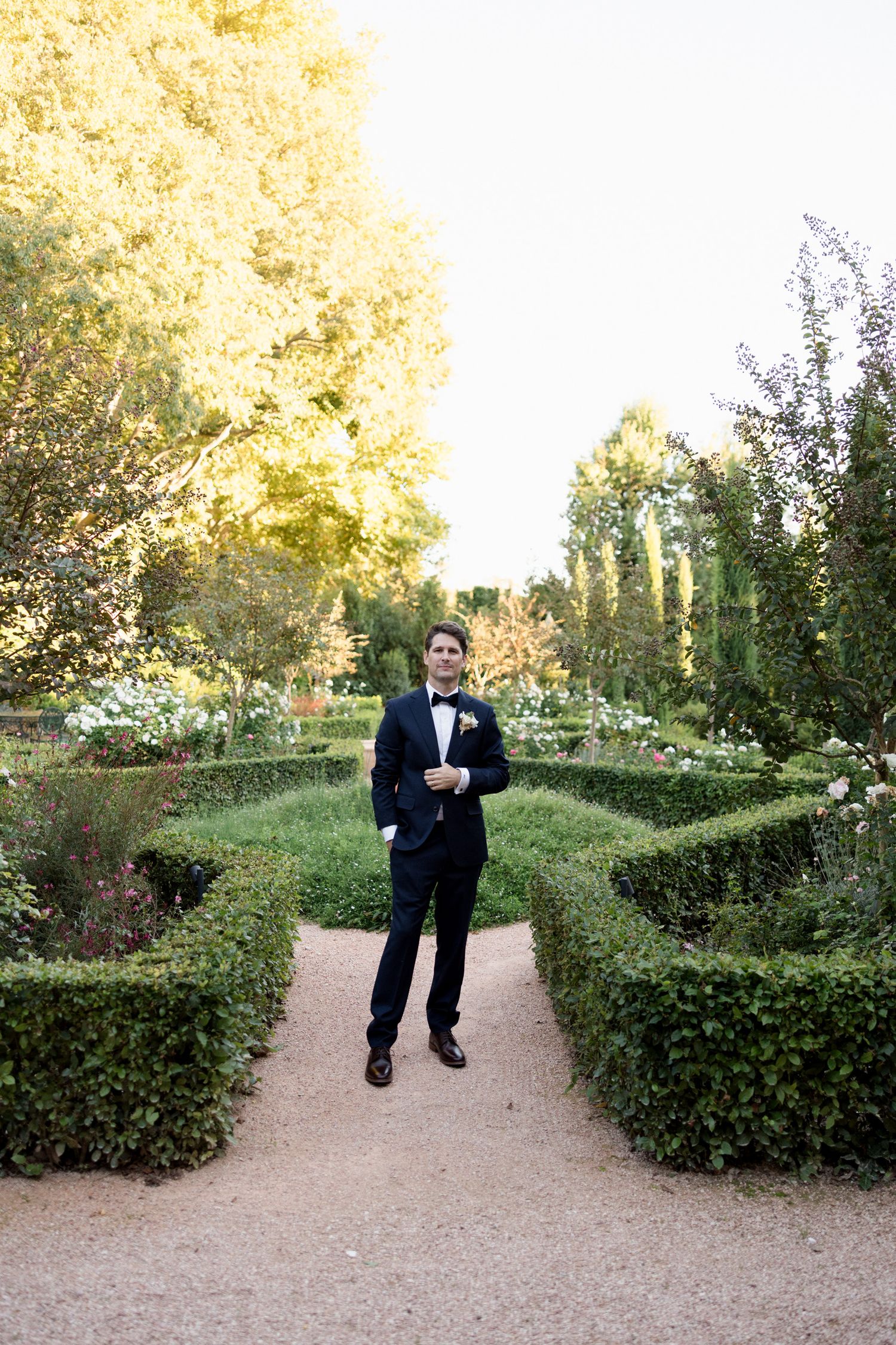 A person in a black tuxedo walks along a winding garden path surrounded by manicured hedges on a sunny day.