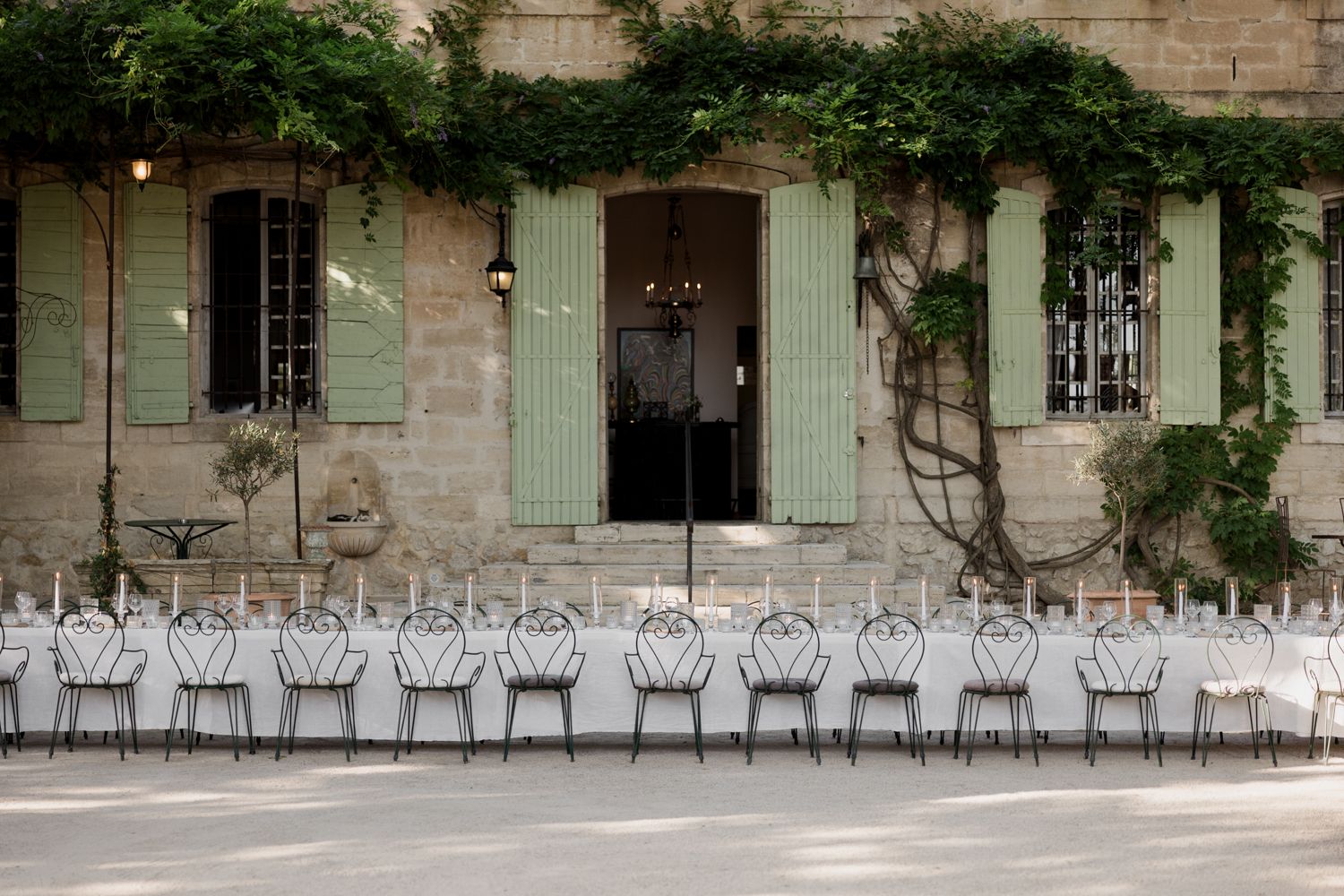 Empty metal chairs lined up outside a rustic French-style building with green shutters and climbing vines. Auberge de la Treille, Avignon