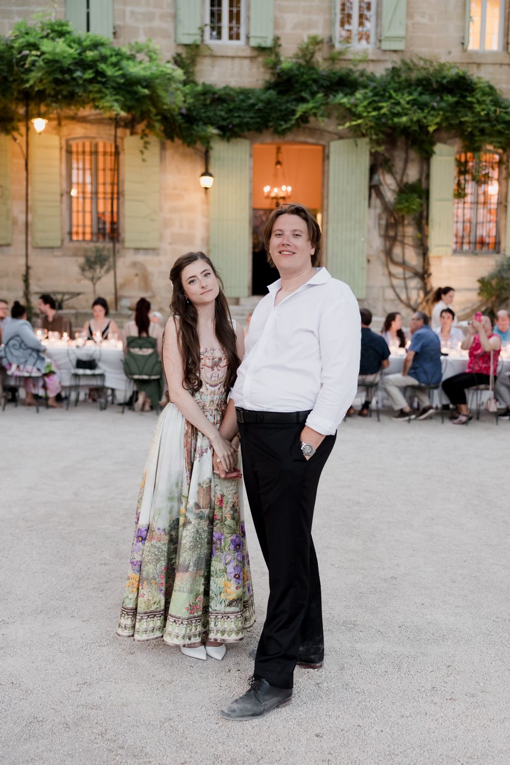 A couple poses in front of l'Auberge de la Treille with climbing vines, as guests dine at outdoor tables in the background. Avignon