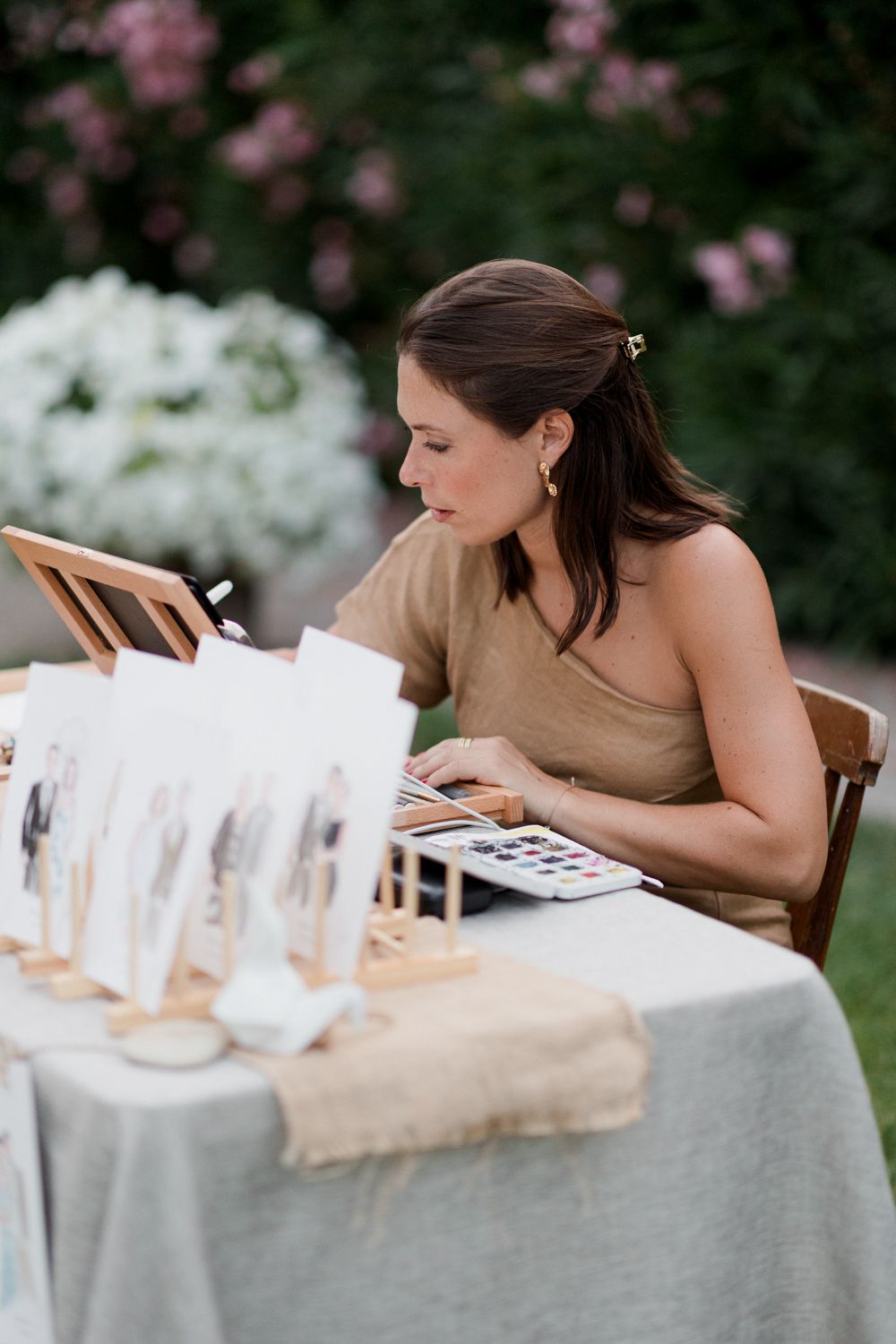 Artist carefully painting watercolor artwork at outdoor table with pink flowers blooming in background. Aurora Artiste