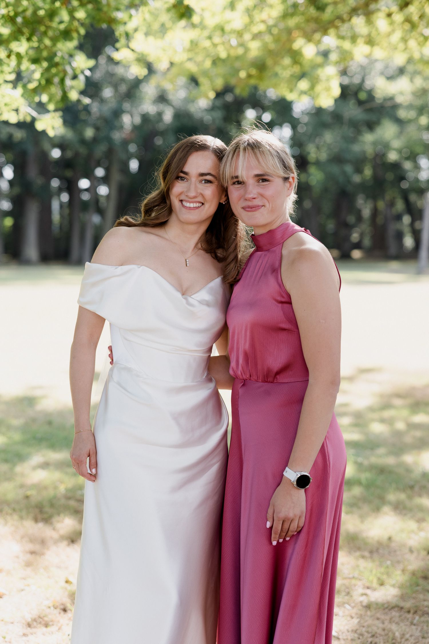 Two people embrace outdoors in formal attire, one wearing a white dress and the other in a dusty rose gown.