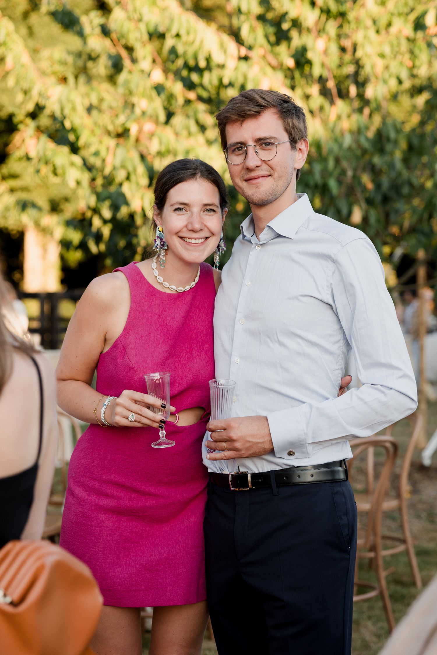 A couple in formal attire holding wine glasses poses together at an outdoor event in warm evening light.