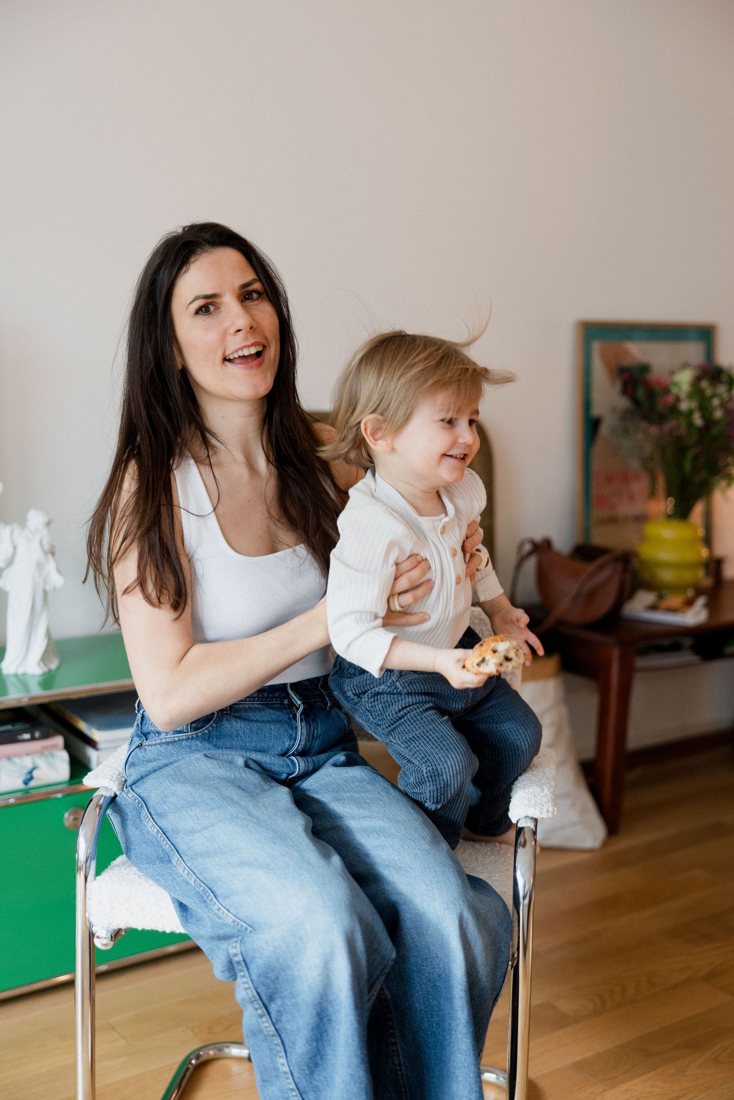 A smiling dark-haired woman in jeans holds a toddler on her lap while sitting on a chair in a cozy home.
