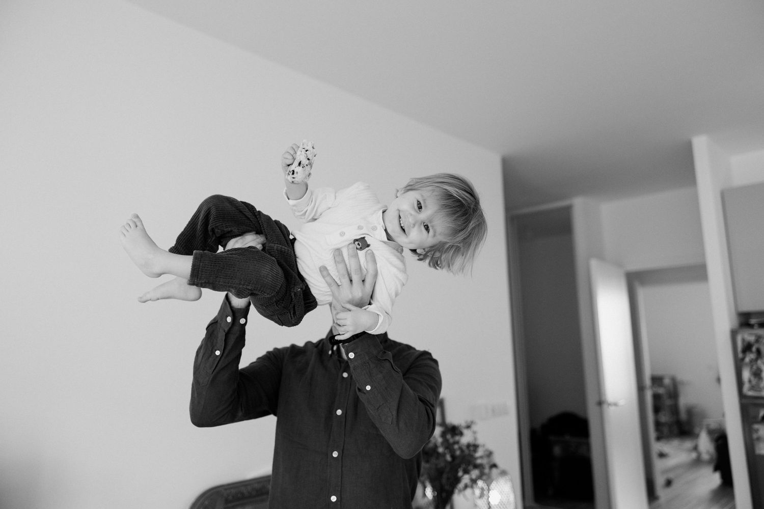 Black and white photo of a father joyfully lifting his laughing toddler high in the air indoors.
