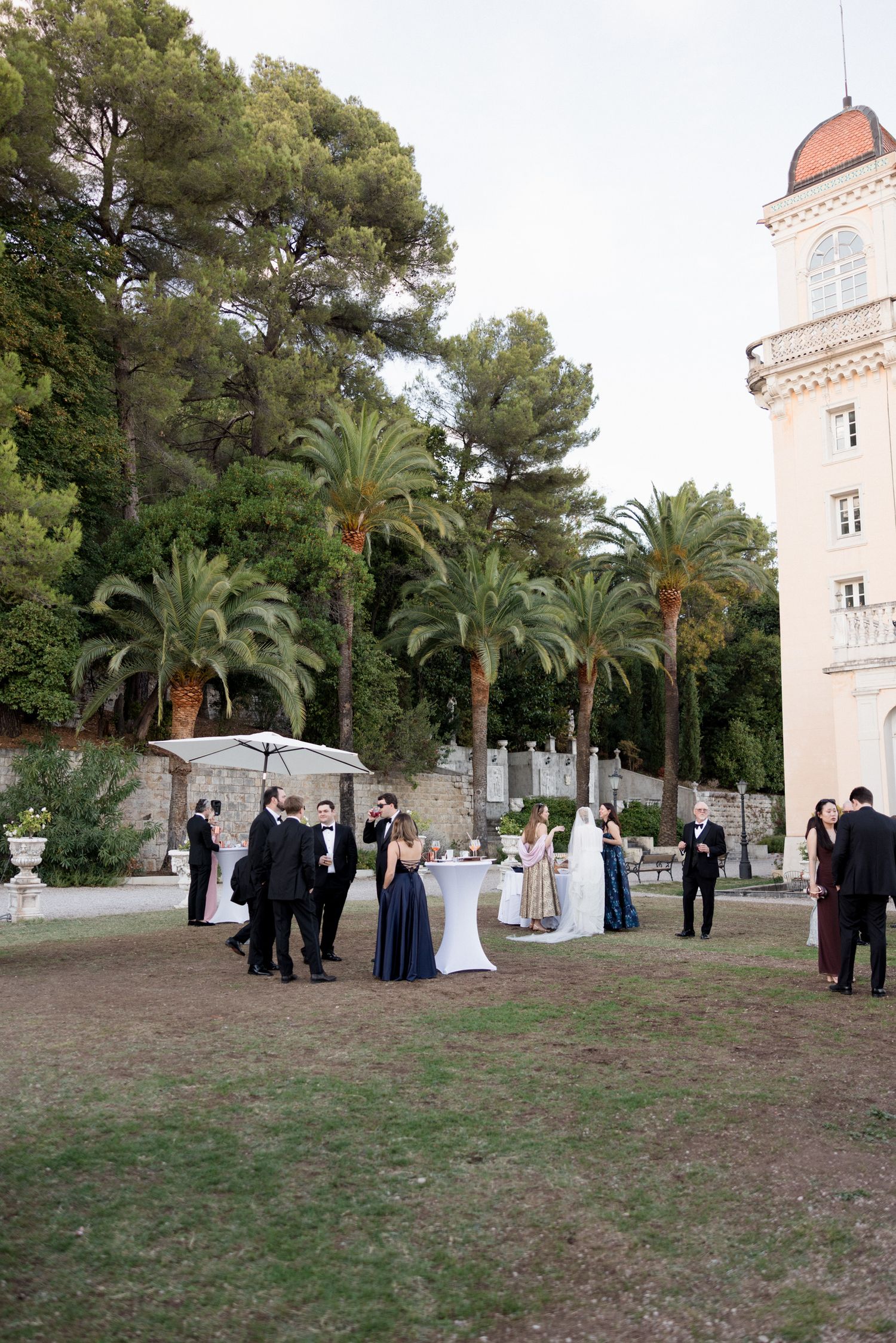 An elegant outdoor wedding reception with guests mingling near palm trees and a historic tower at dusk.