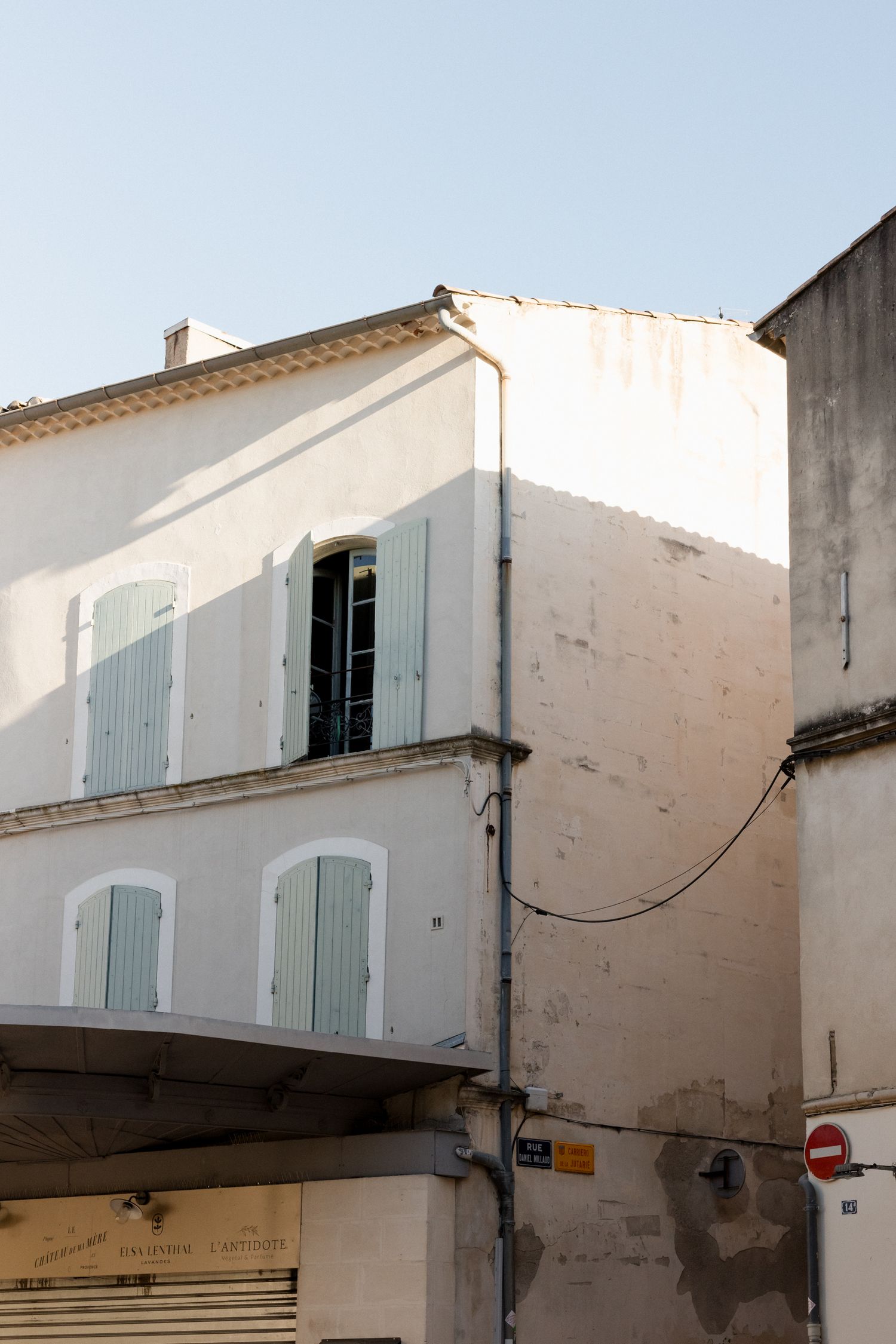 Mediterranean style buildings with white walls and pale blue shutters line a narrow street in a quaint village setting.
