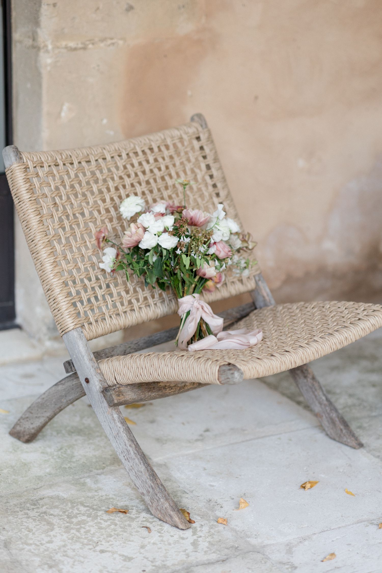 A rustic woven rope chair with weathered wooden frame holds a delicate bouquet of white flowers against a textured wall. wedding photographe